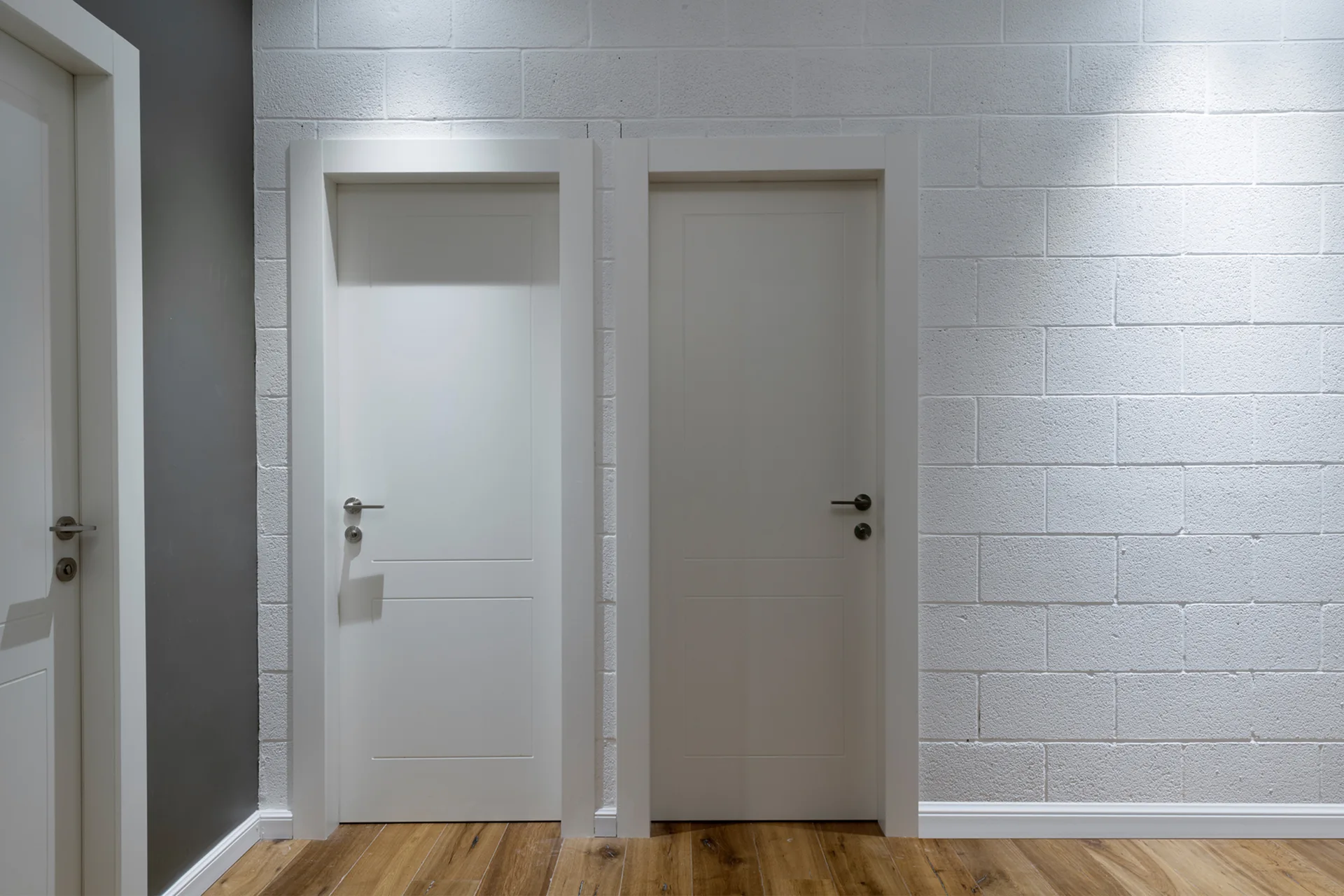 Two white panel doors set in a painted white cinder block wall in a hallway with warm wood floors.