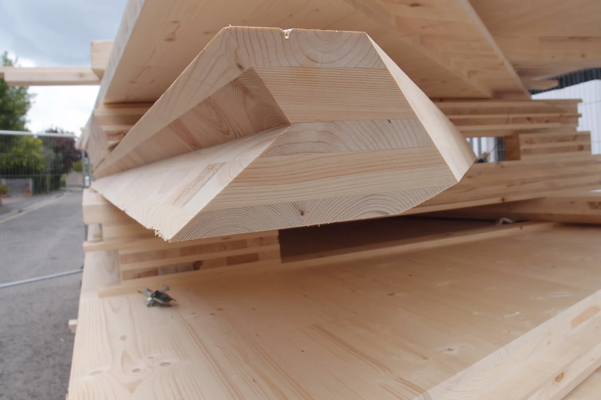 A stack of pale laminated timber beams shows layered wood grain and angled profiles in an outdoor construction setting.