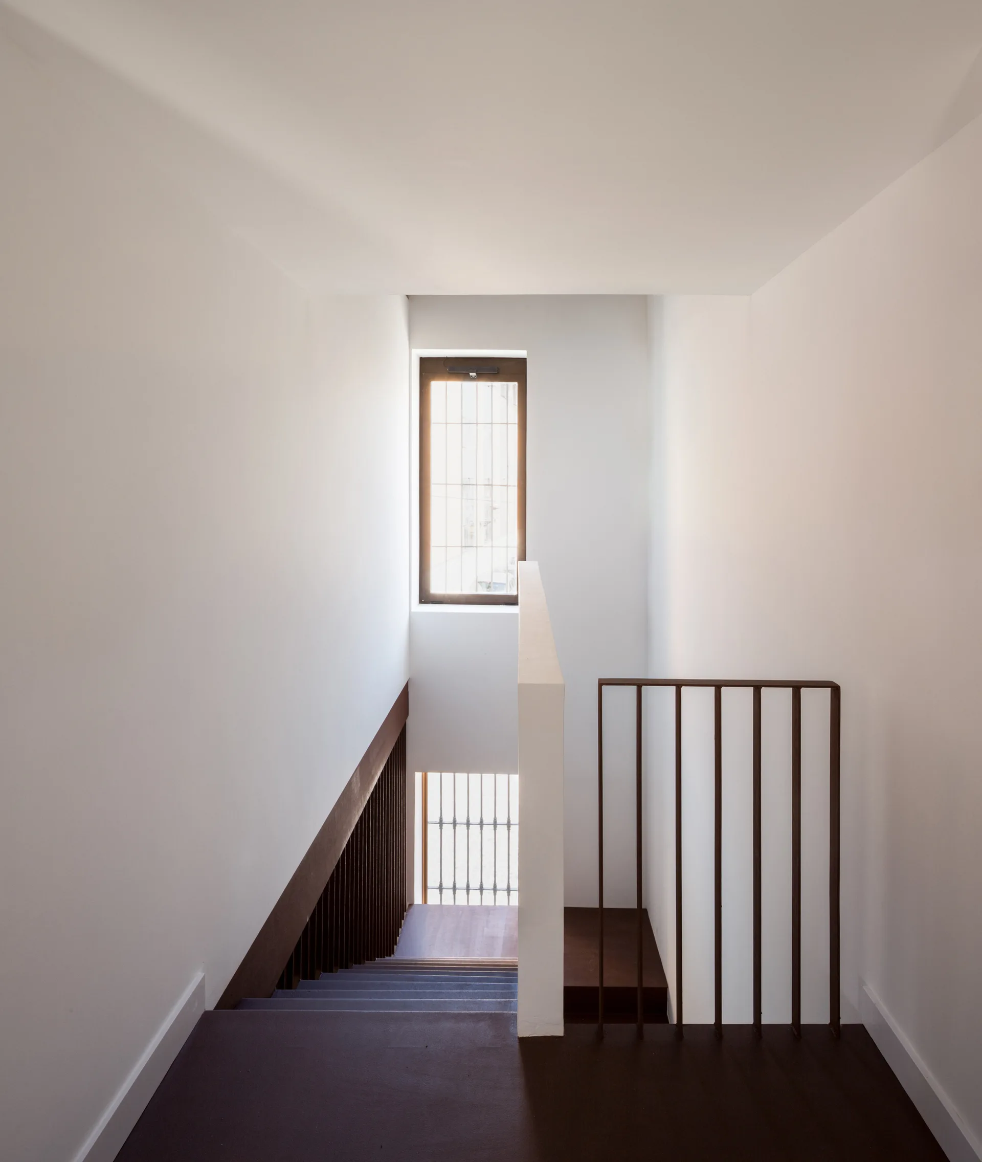 A minimalist stairwell features dark brown wooden steps, slim vertical metal railings, and a rectangular window set in smooth white walls.