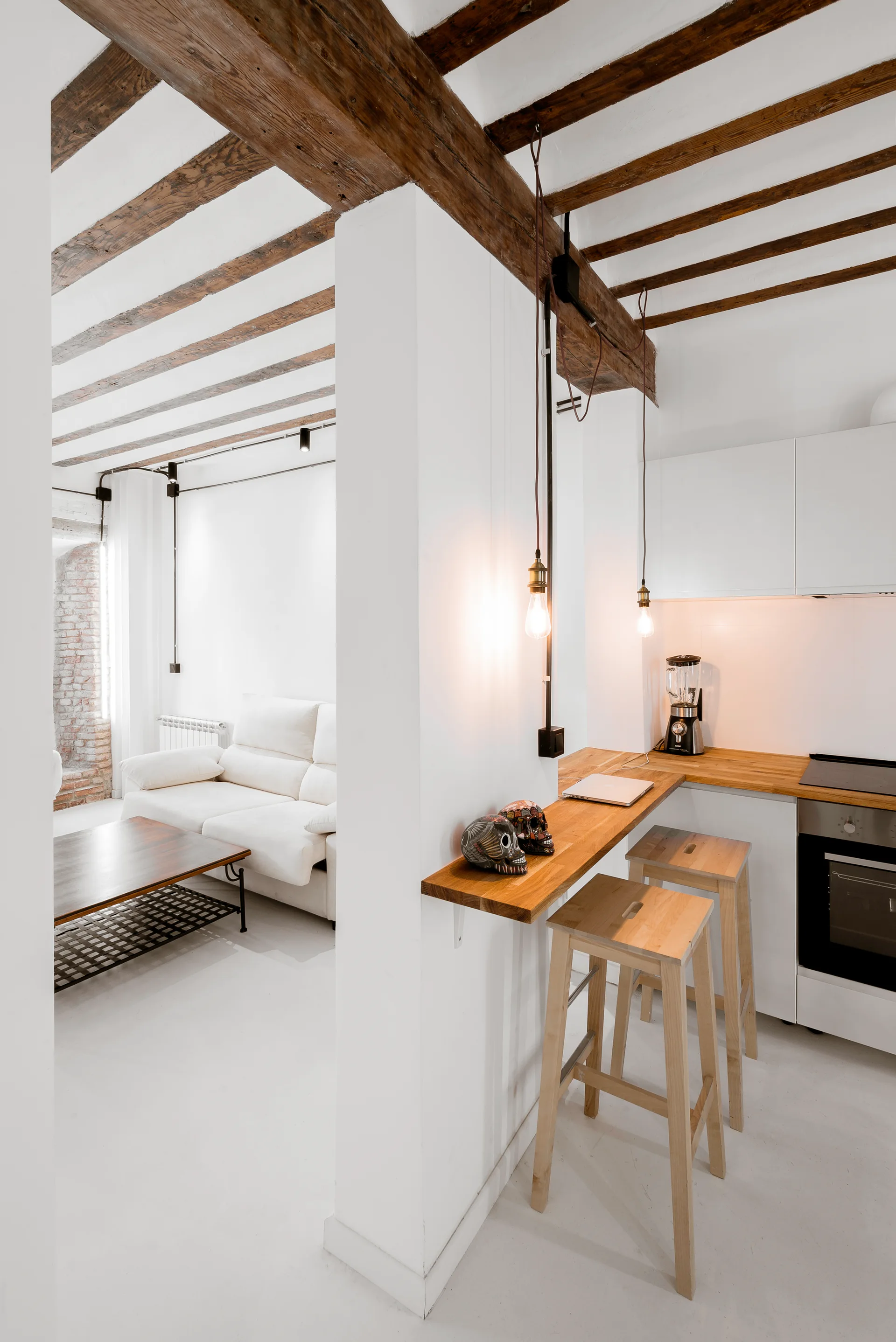 A white kitchen features a wooden breakfast bar with bar stools below rustic ceiling beams next to a living area.