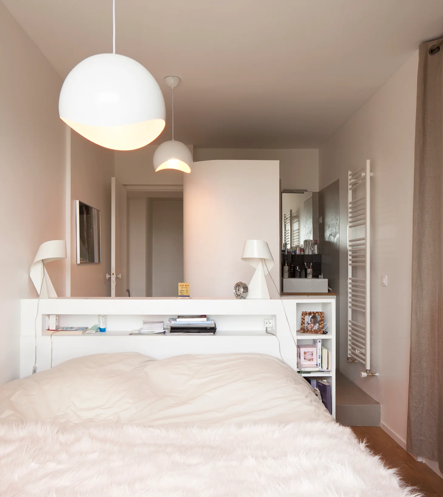 A minimalist white bedroom featuring a bed with an integrated headboard shelf divider, hanging dome pendant lights, and a sink area in the background.