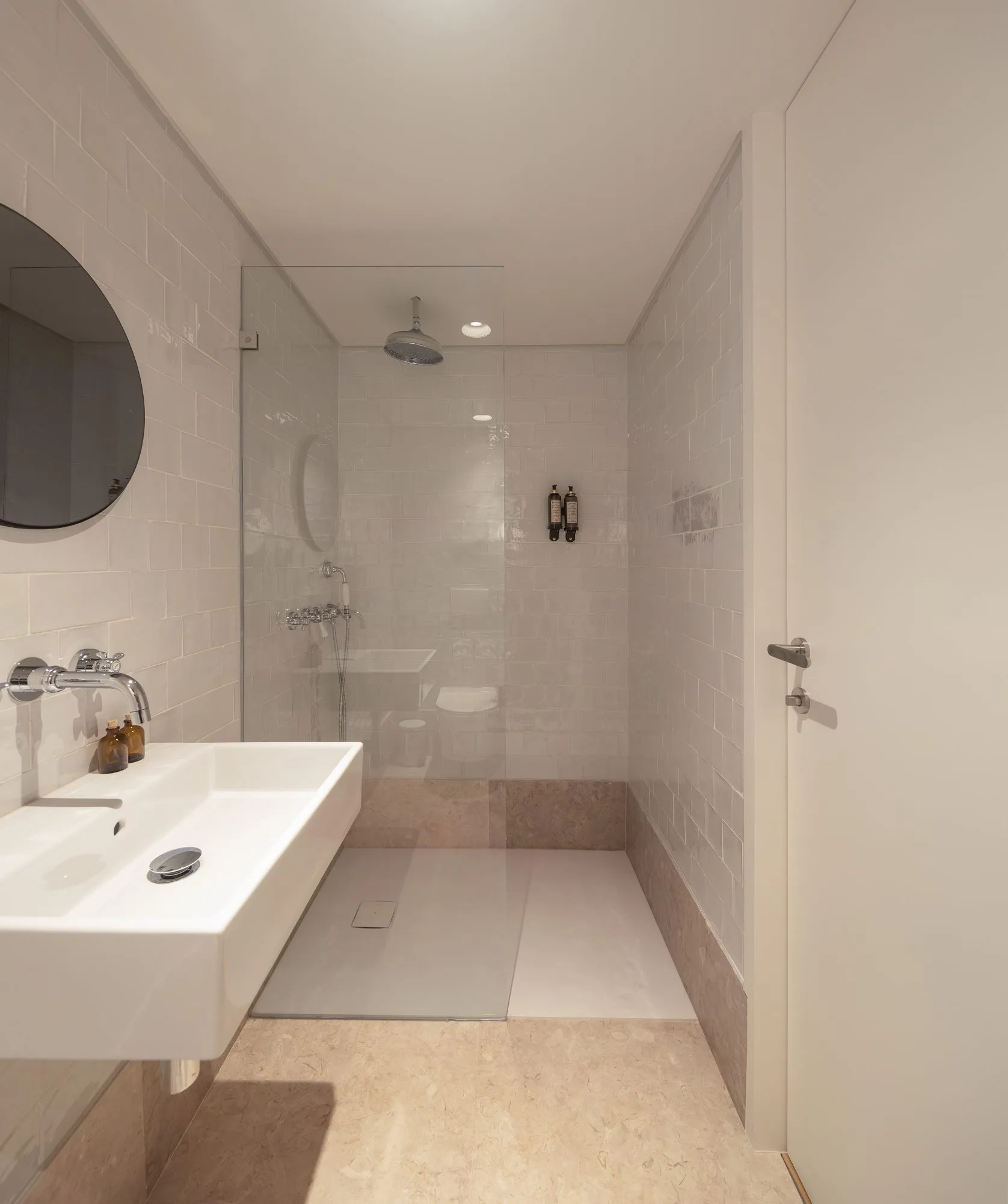 A modern bathroom features a wall-mounted white sink, a frameless glass walk-in shower, glossy white subway tiles, and beige stone flooring.