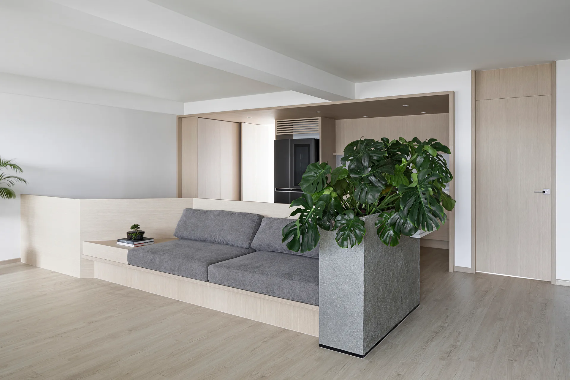 A minimalist living room features a grey cushioned sofa built into a light wood platform beside a tall textured grey planter with a Monstera plant.