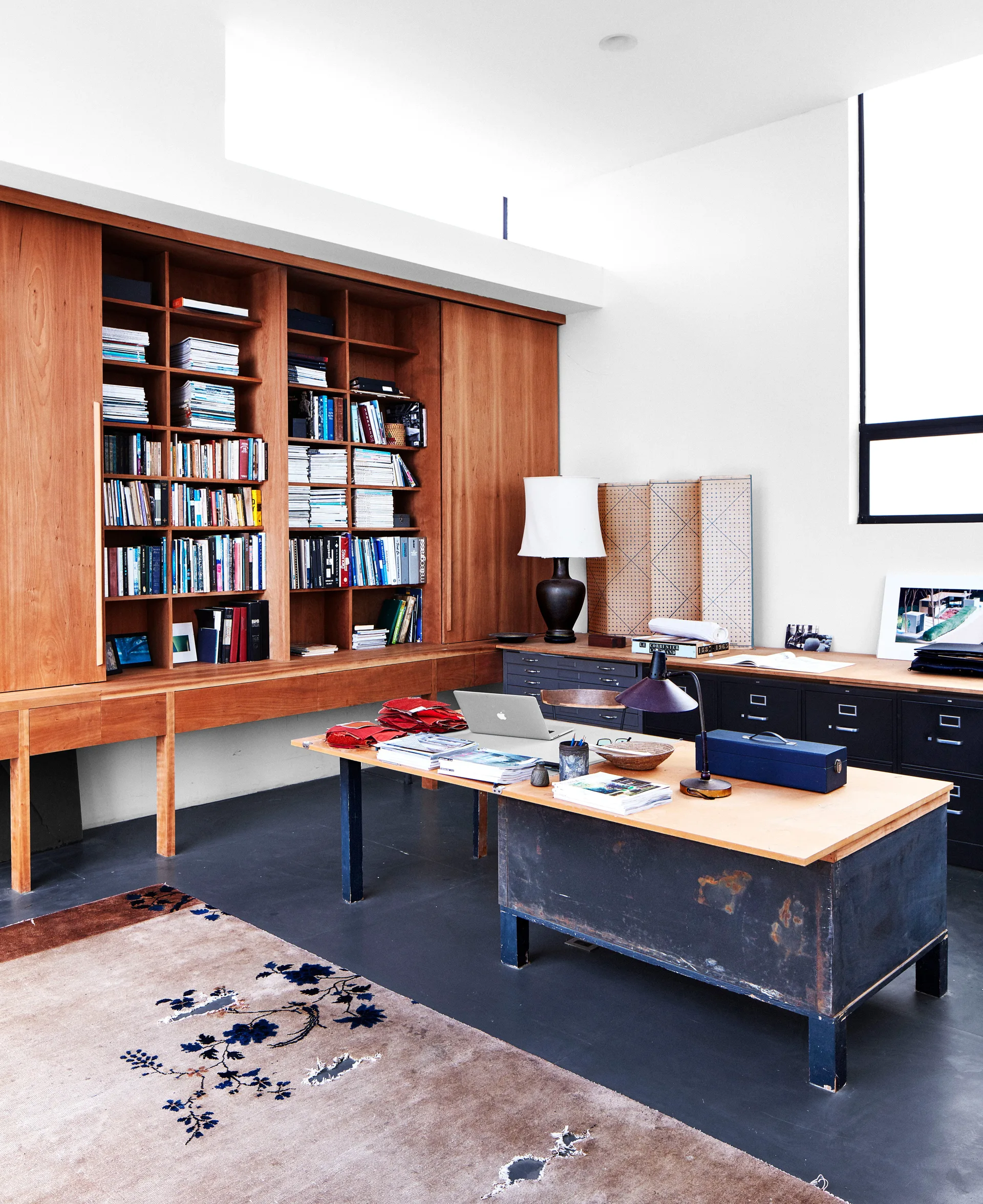 An industrial style home office features a large built-in wooden bookcase, a metal desk with a wood top, black file cabinets, and a distressed area rug.