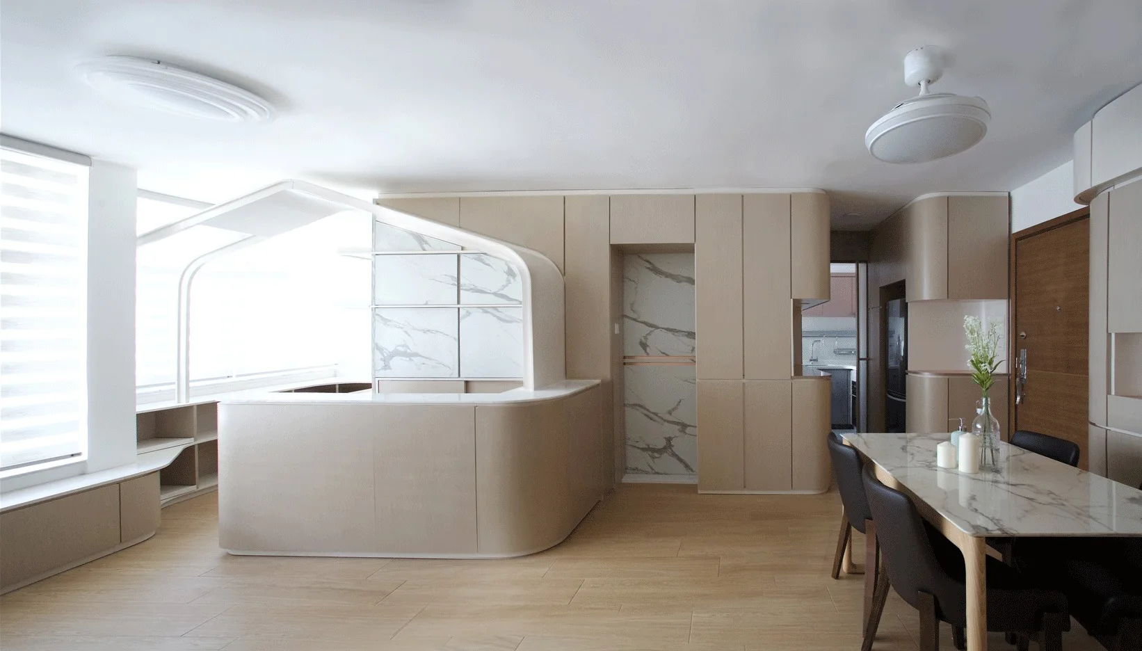 A modern dining space features custom beige curved cabinetry, a marble-topped table with dark chairs, and light wood floors.