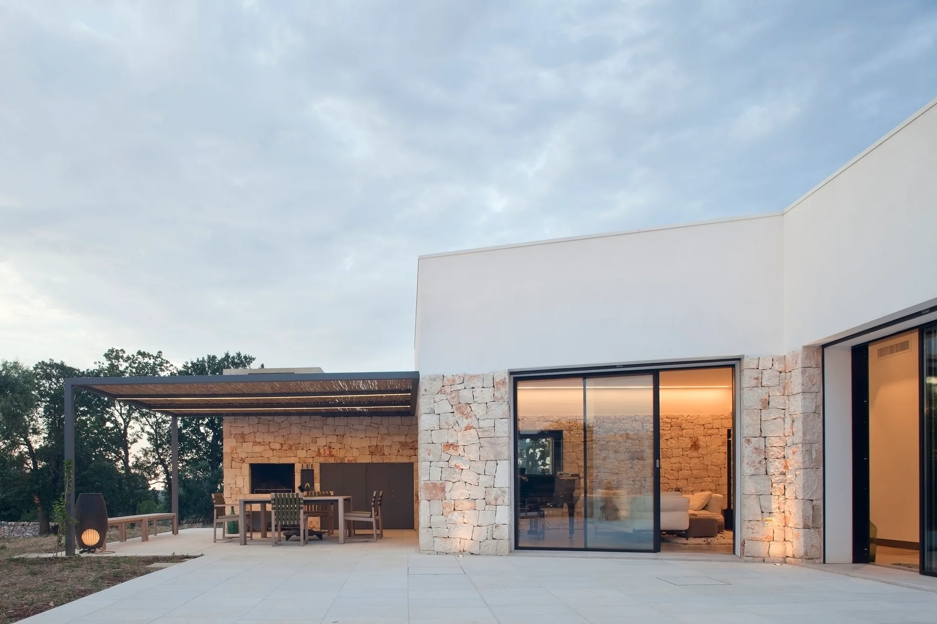 A modern paved patio features a rough stone wall facade, a slatted pergola over a dining set, and large sliding glass doors revealing a warm interior.