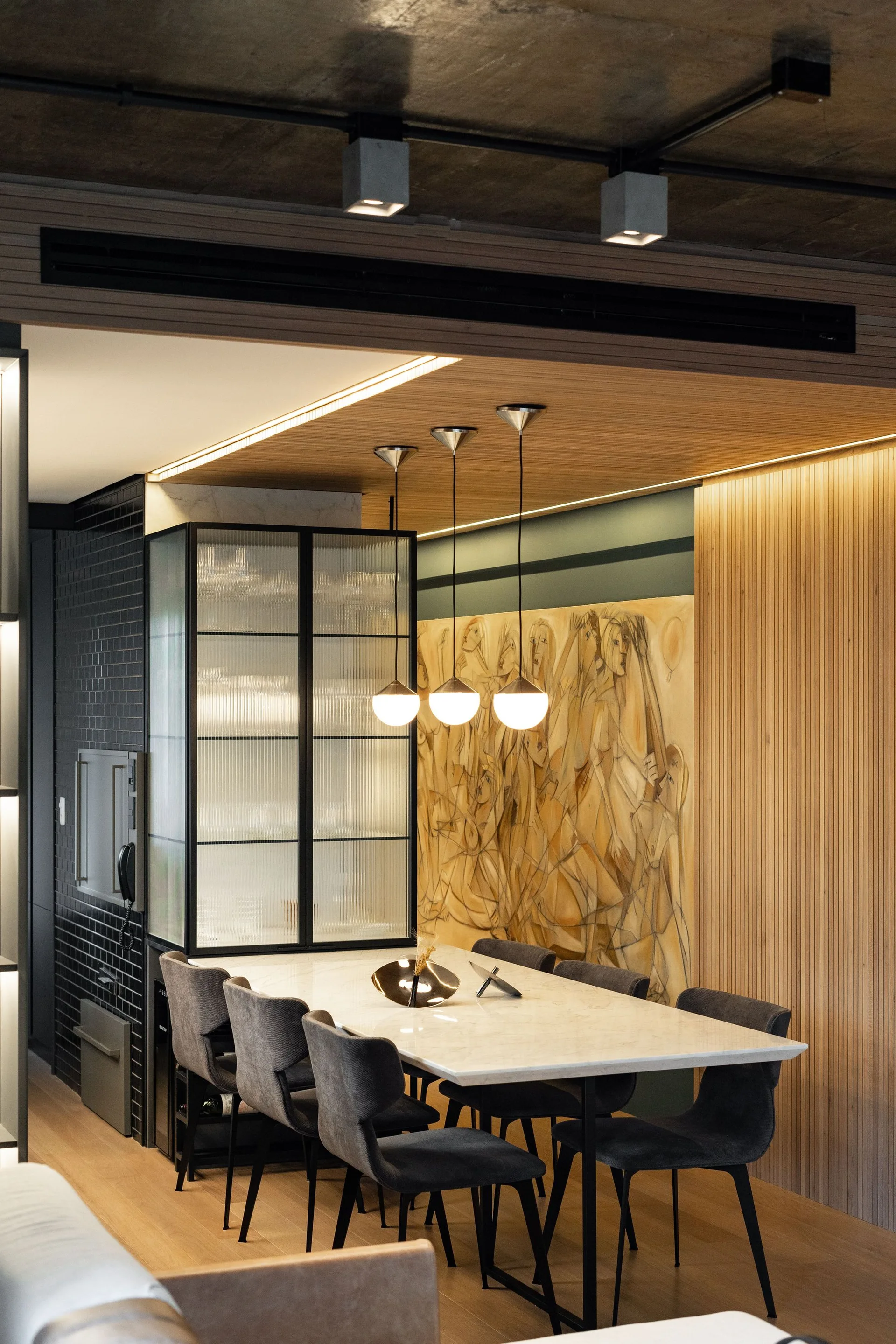A contemporary dining room features a marble table with grey chairs beneath globe pendants, next to a wood slatted wall and glass cabinet.