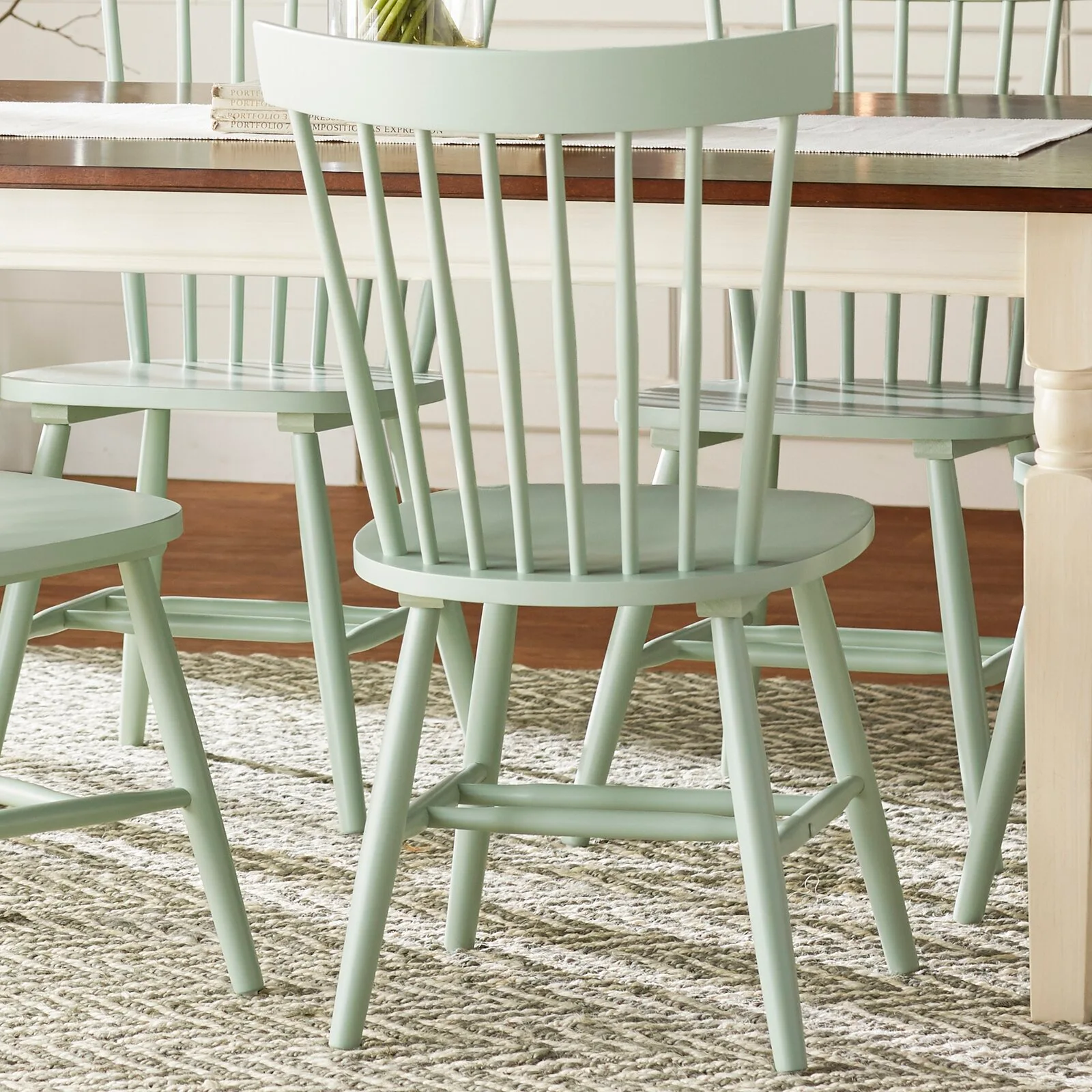 Sage green spindle-back chairs are arranged around a wooden dining table on top of a textured neutral rug.