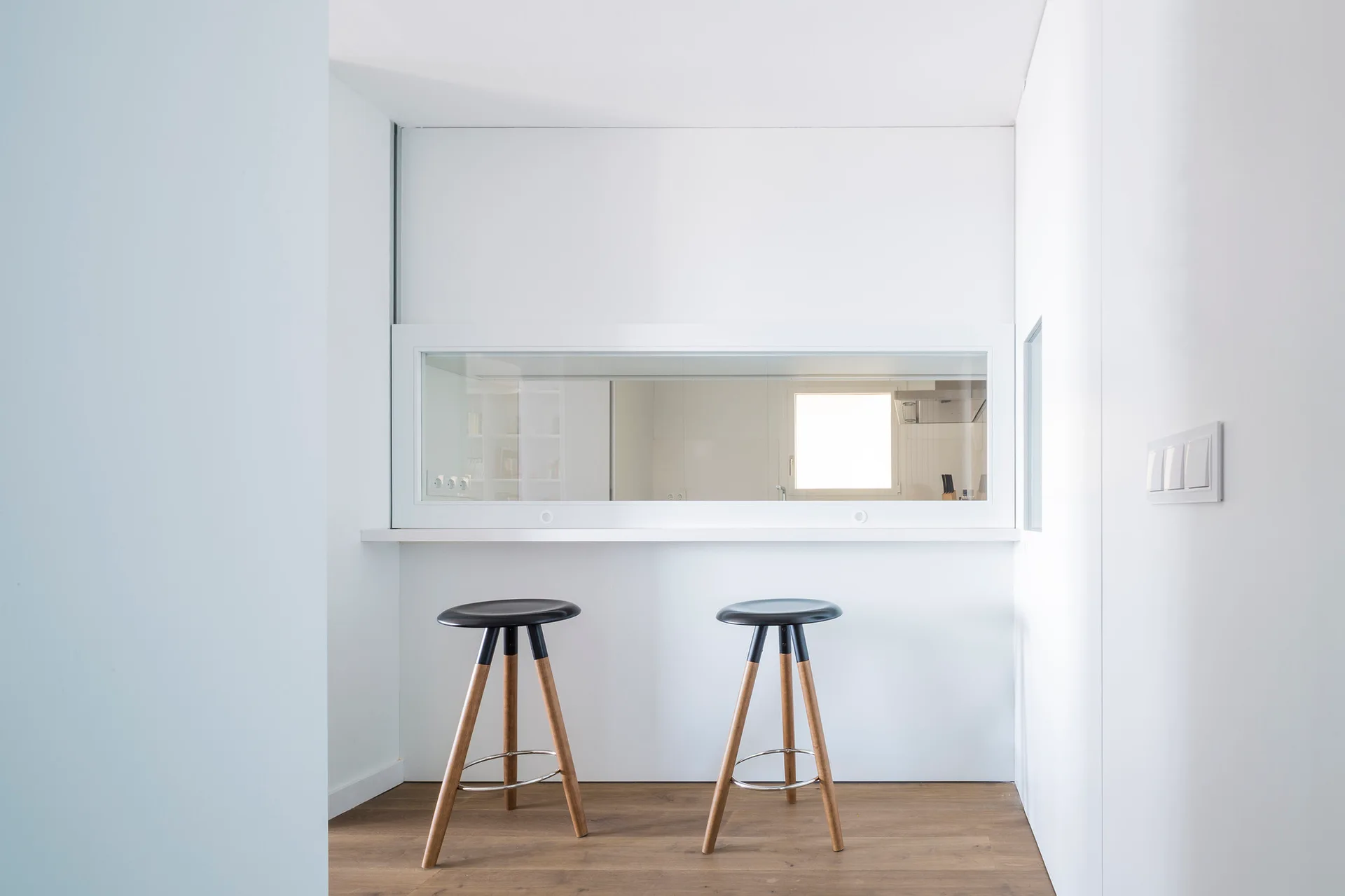 Two minimalist bar stools with wooden legs and black seats stand at a white breakfast bar beneath a wide pass-through kitchen window.