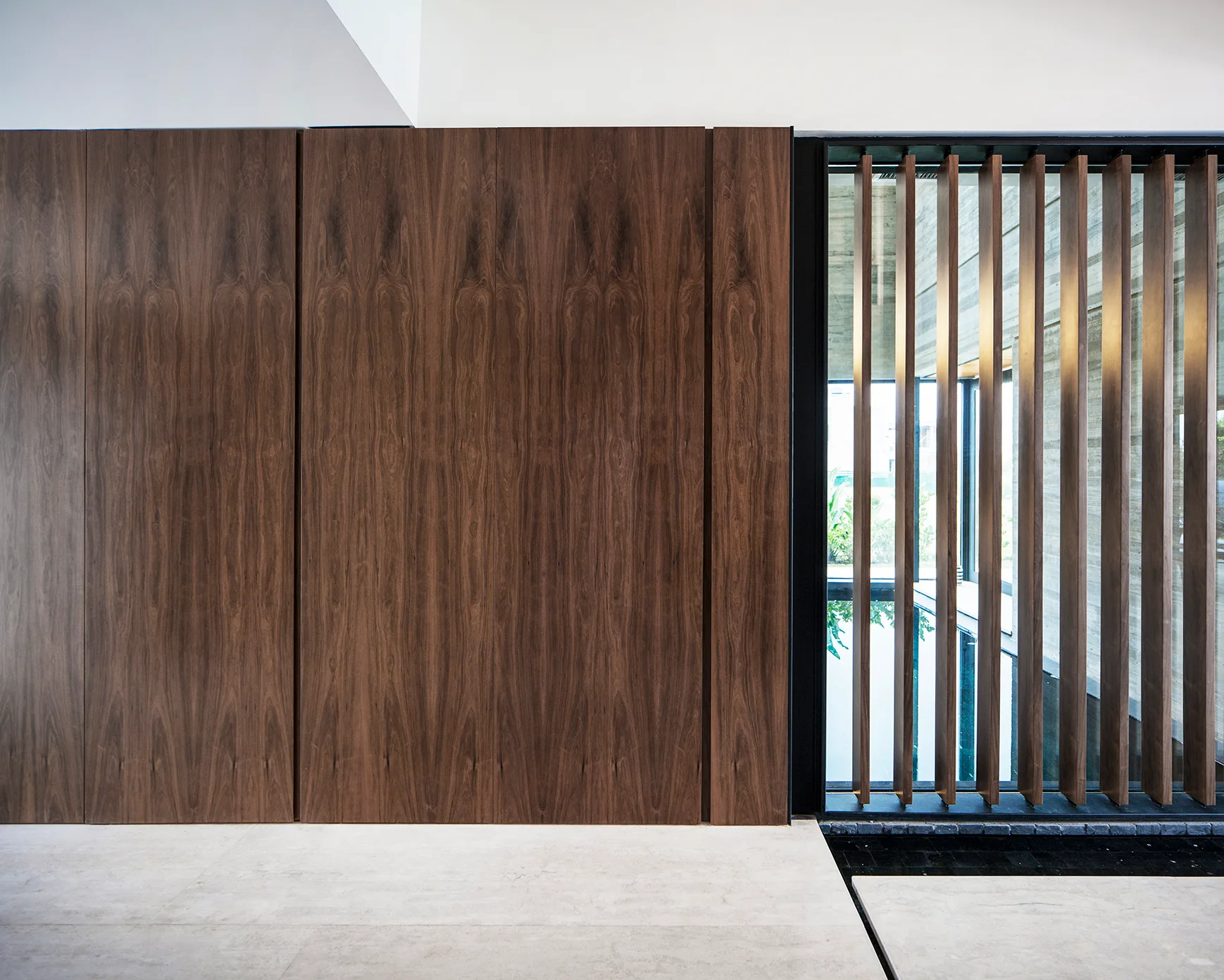A minimalist interior wall features seamless brown wood paneling next to a tall window with vertical wooden slats over a light stone tile floor.