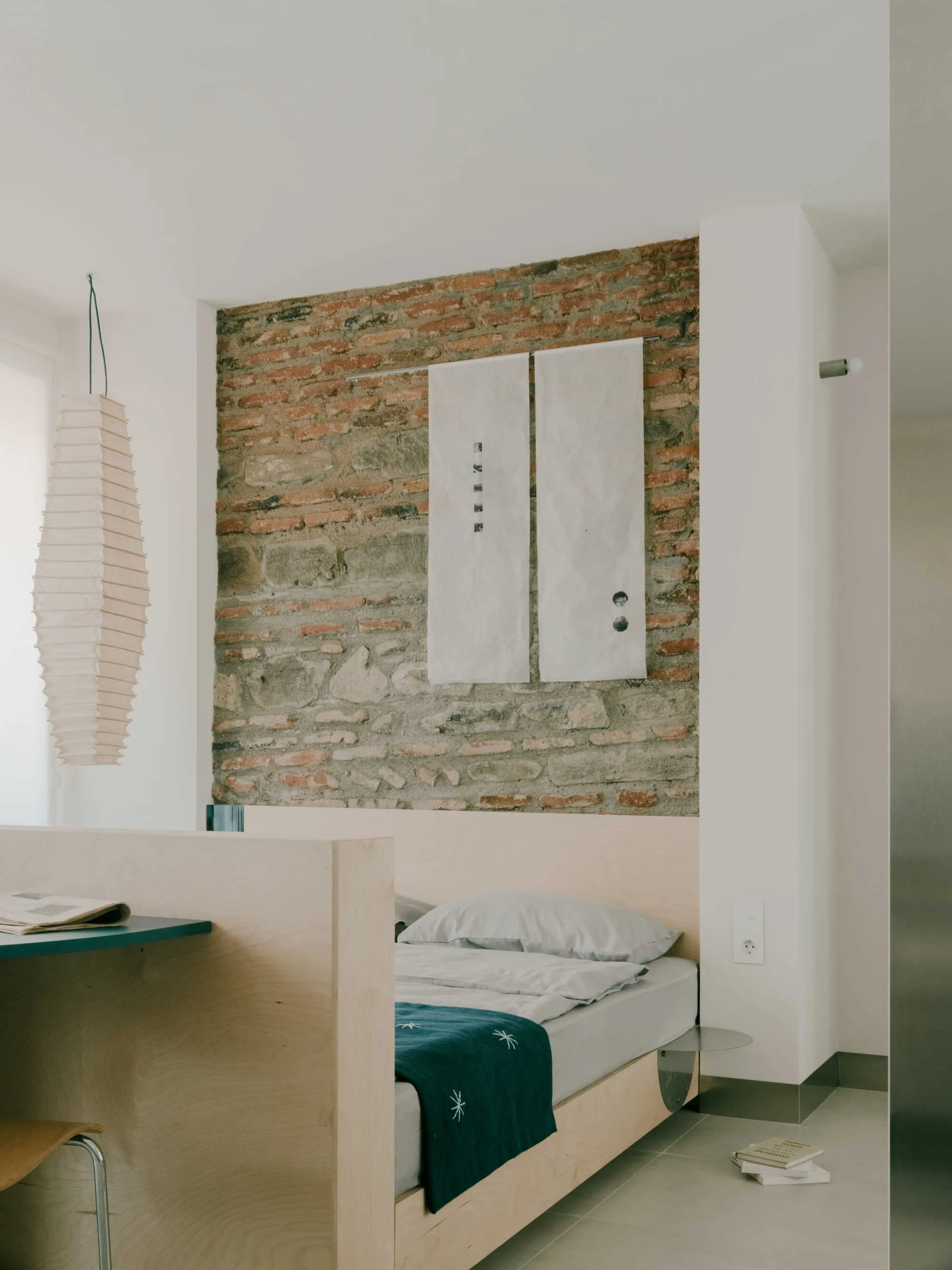 A minimalist bedroom features a light wood platform bed with a wooden headboard partition against a rustic exposed brick and stone wall.