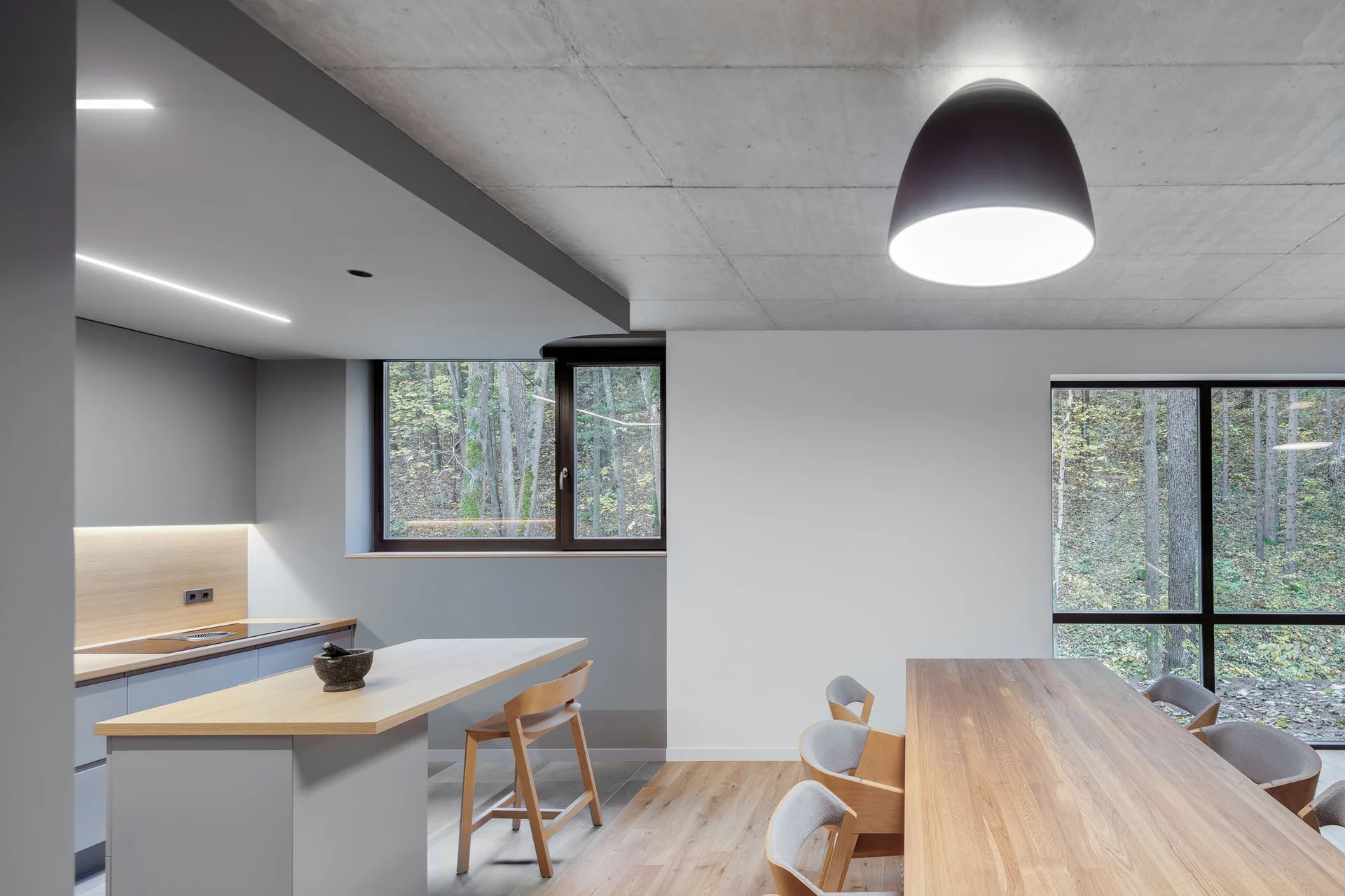 A minimalist dining area and kitchen with an exposed concrete ceiling, a light wood table, and large windows looking out at trees.