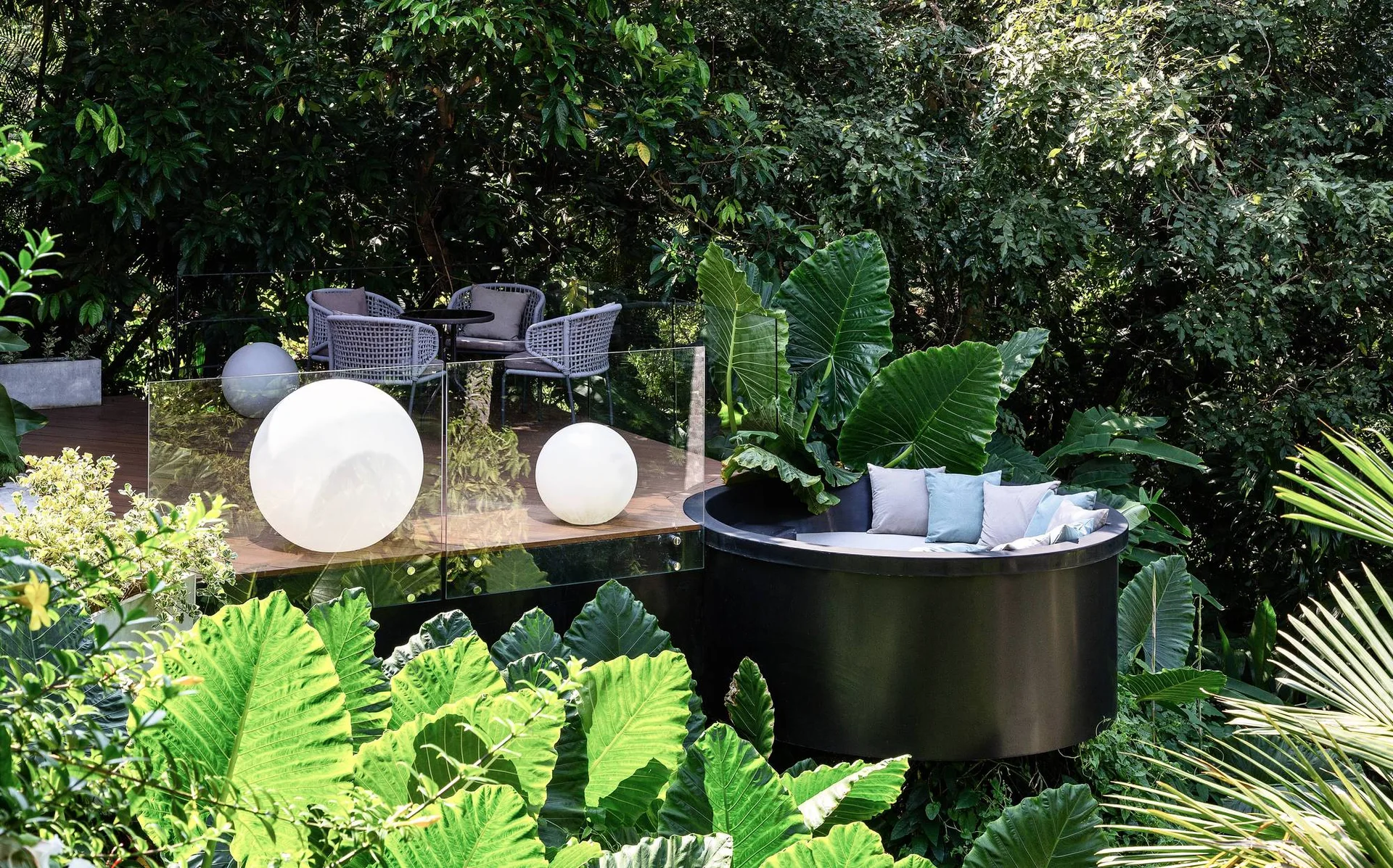 A modern wooden deck featuring a glass railing, glowing orb lights, woven chairs, and a black circular daybed surrounded by dense tropical foliage.