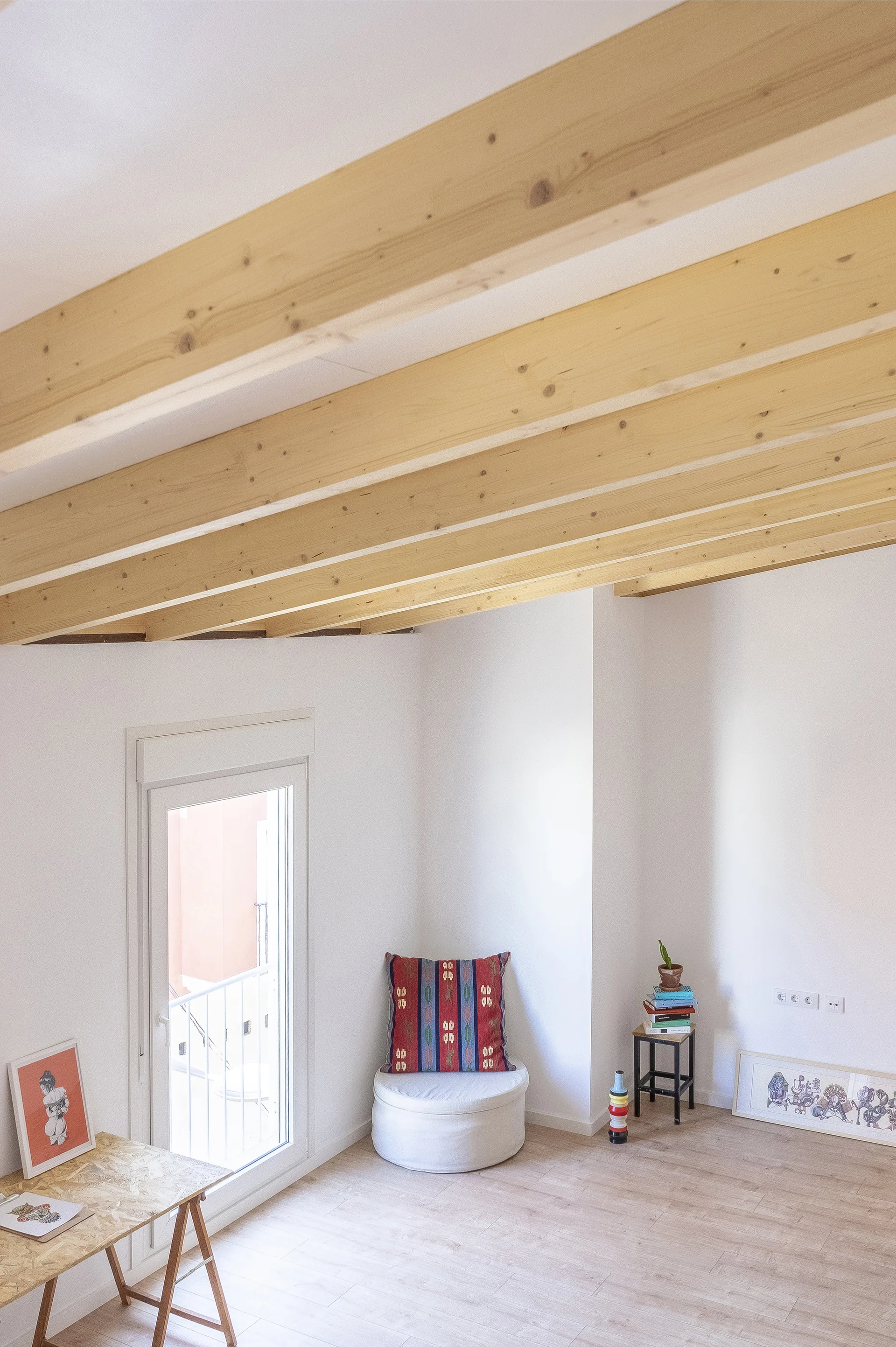 A minimalist room features exposed pine ceiling beams, light wood floors, a white corner ottoman, and a tall glass door.