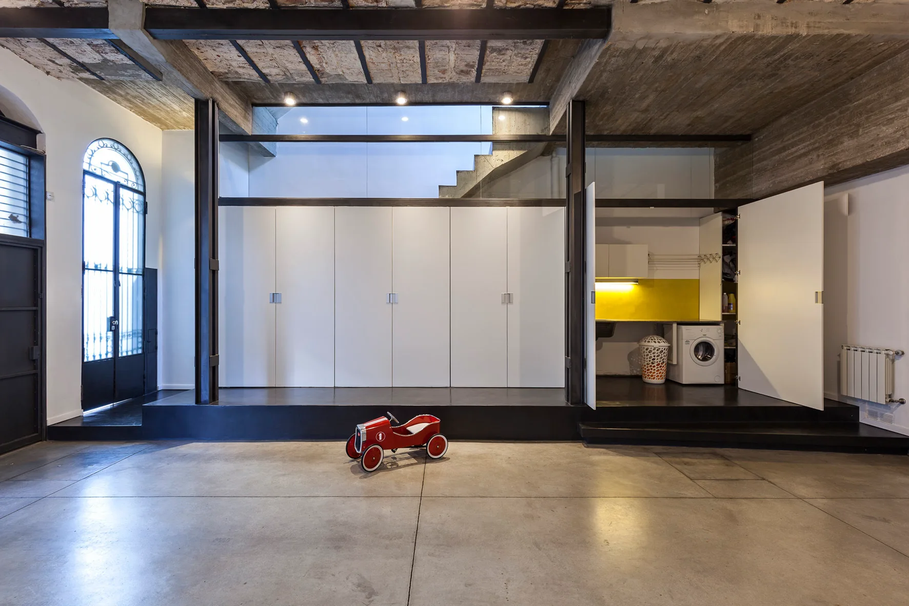 An industrial space featuring a raised black platform with white built-in storage cabinets, one of which opens to reveal a laundry washer nook.