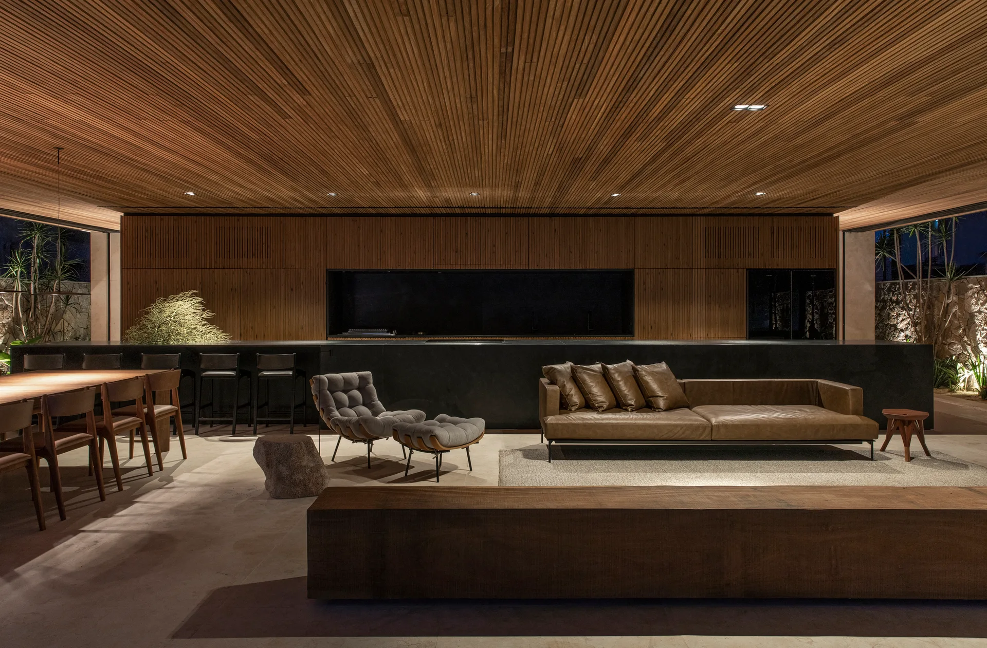 A modern open-plan living room features a brown leather sofa, a long wood-slat ceiling, integrated wood-paneled walls, and a large black island.