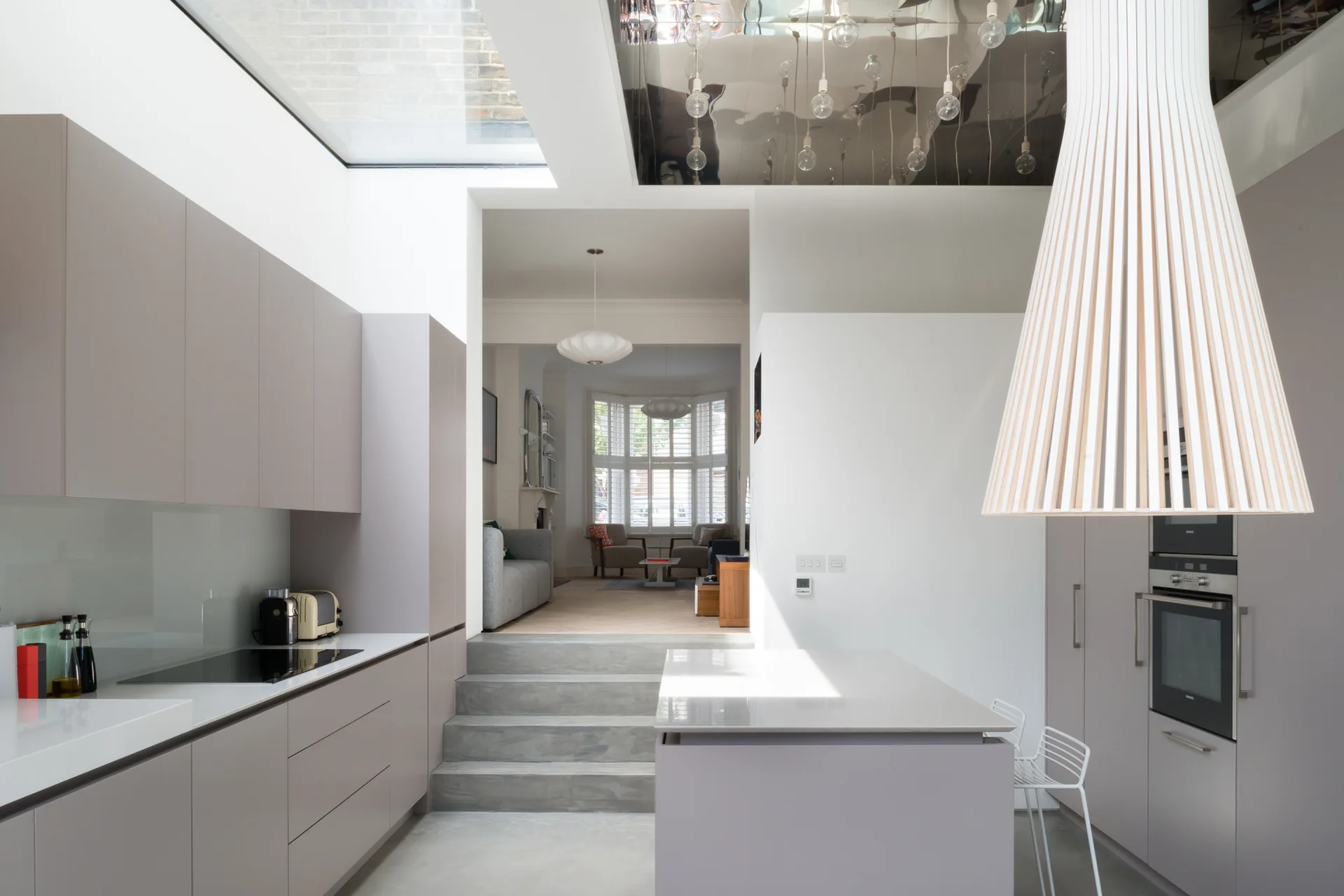 A modern split-level kitchen with grey handleless cabinets, a concrete floor, and steps leading to an upper living room under a bright skylight.