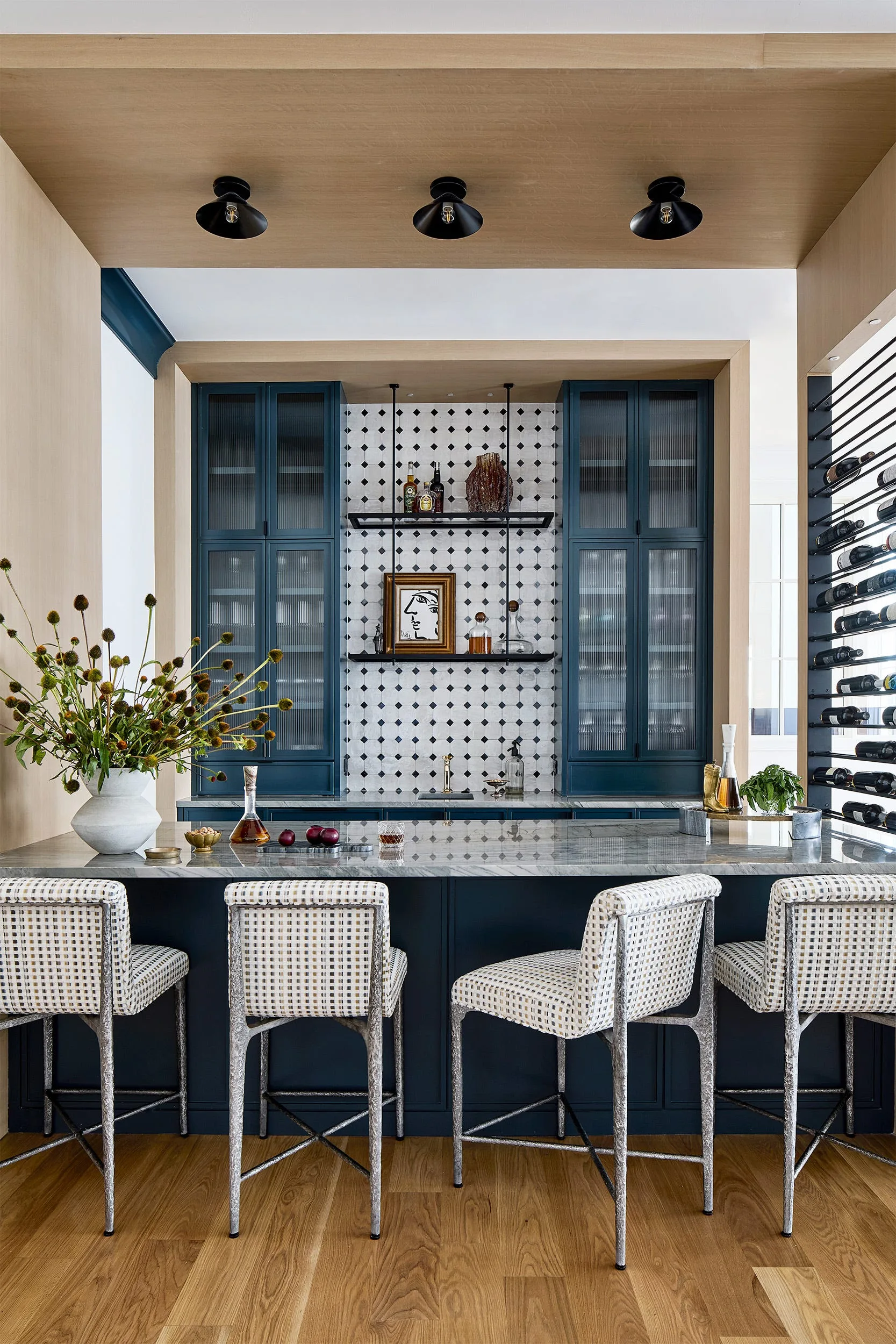 A home bar features navy blue cabinets with reeded glass doors, a patterned tile backsplash, a marble countertop, and four upholstered bar stools.
