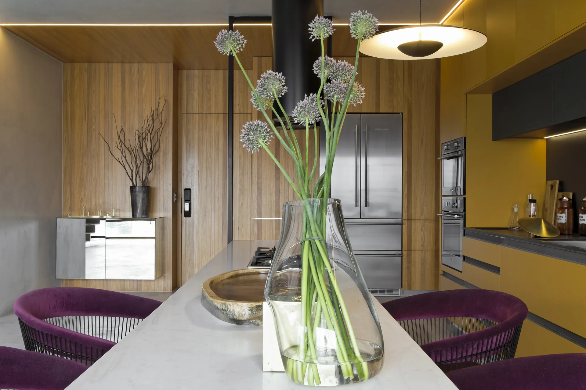 A modern kitchen featuring a stone-topped island with purple velvet chairs, set against vertical wood-slat walls and bold yellow cabinetry.