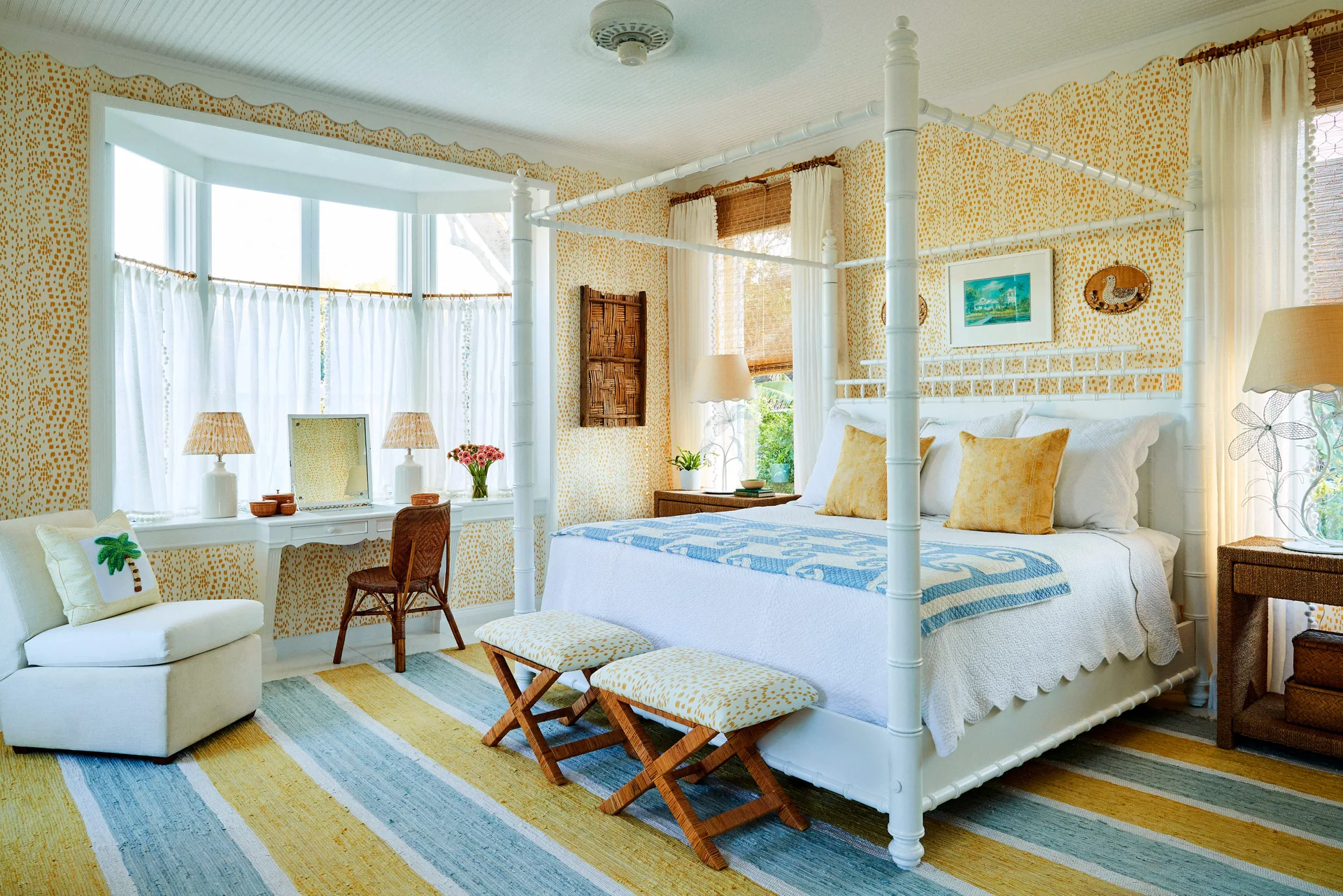 A bright cottage bedroom featuring a white four-poster bed, yellow dotted wallpaper, and a vanity table tucked into a large bay window.