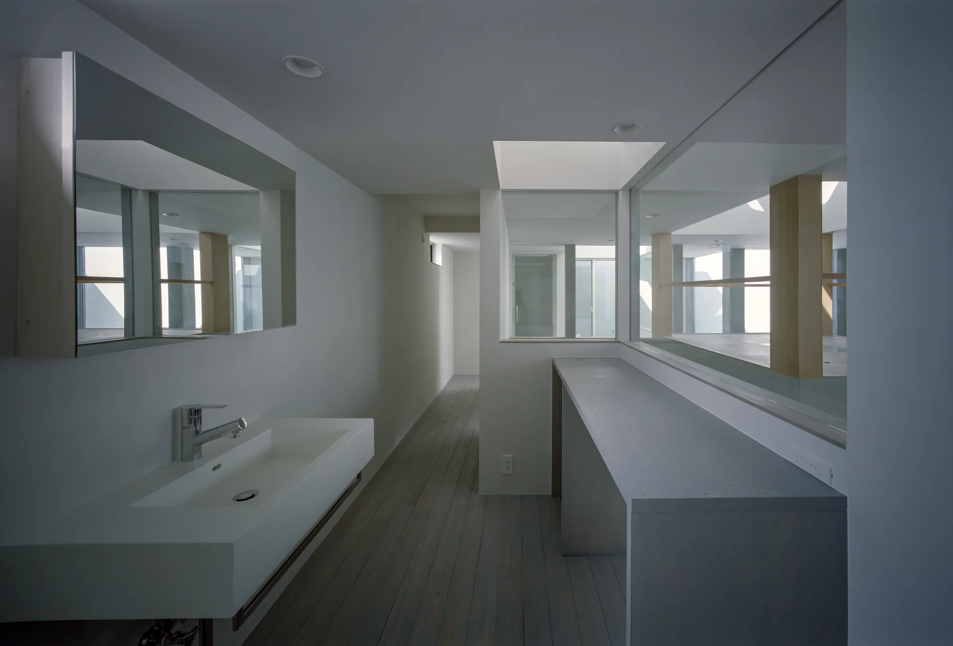 A minimalist bathroom hallway features a wall-mounted white trough sink, a large rectangular mirror, and an internal glass window overlooking another room.