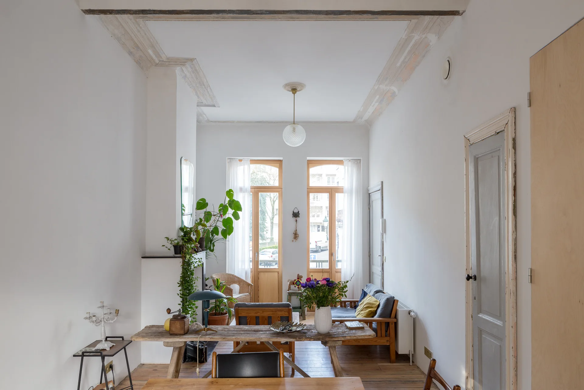 A long, bright living and dining room features a rustic wooden table, a grey sofa, and tall wood-framed French windows under a high decorative ceiling.