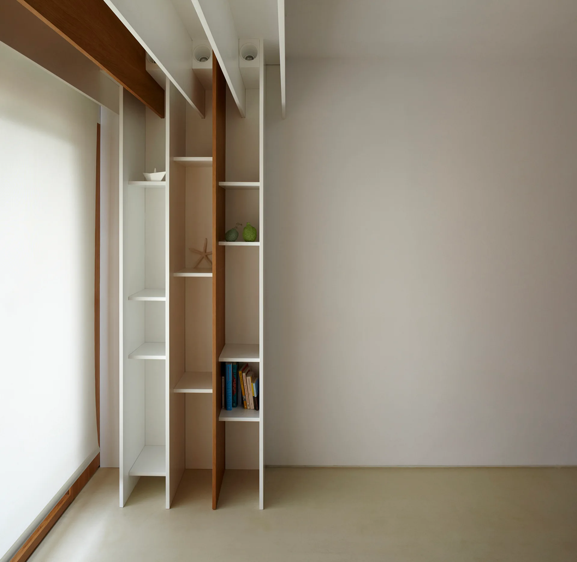 A floor-to-ceiling minimalist shelving unit in white and natural wood integrated into white ceiling beams next to a bright window with a roller shade.
