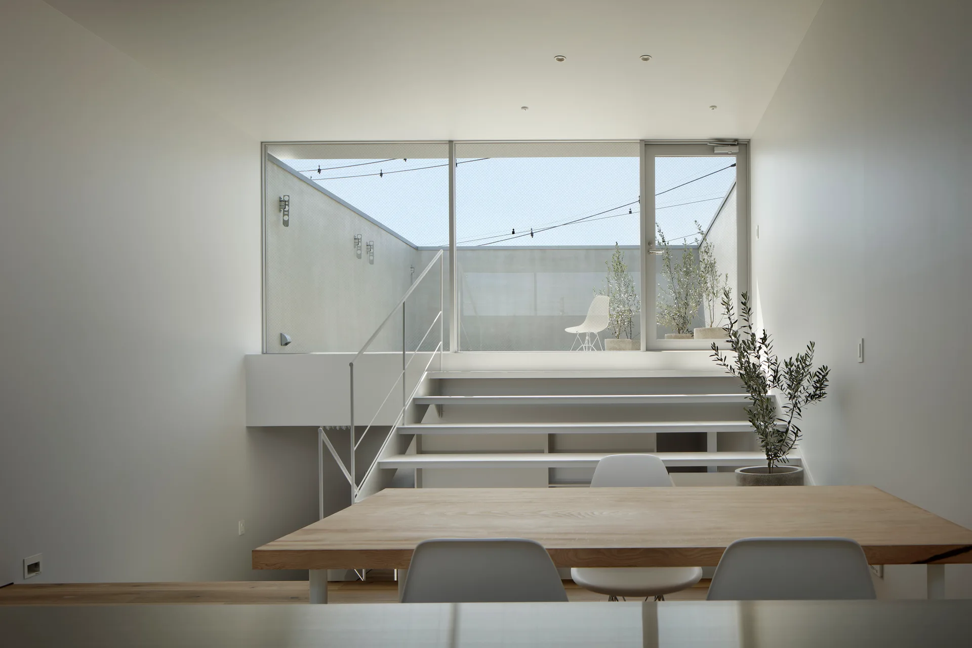 A minimalist dining room features a light wood table, white chairs, and stairs leading up to an elevated terrace behind a large glass window.