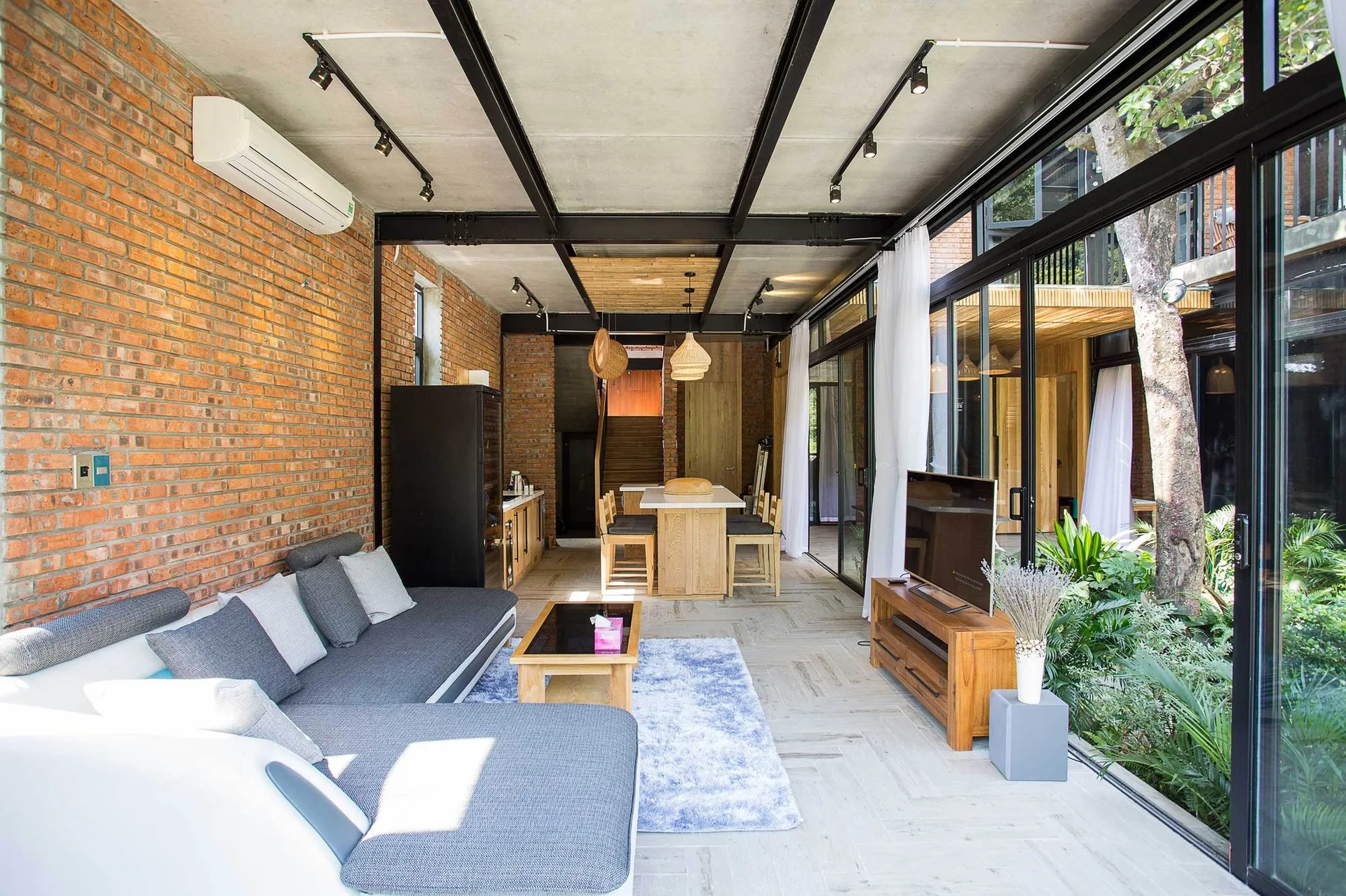 An open-plan industrial living room features an exposed brick wall, a grey sectional sofa, and floor-to-ceiling glass sliding doors opening to plants.