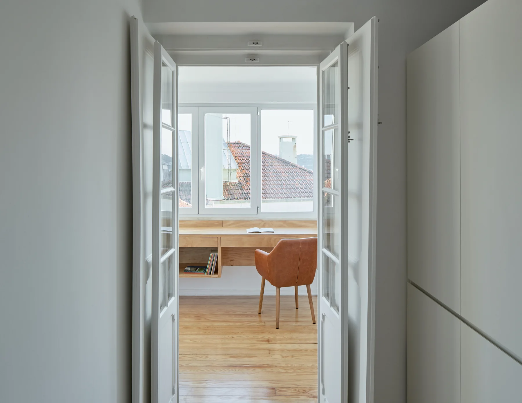 A view through open white French doors into a bright home office featuring a built-in light wood desk under a window and a tan leather chair.