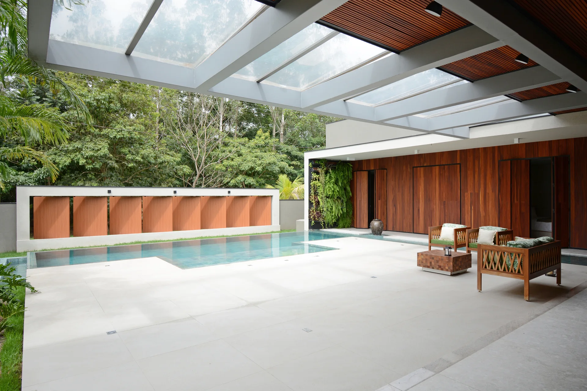 An outdoor patio with a swimming pool, wooden paneling on the walls, a vertical garden, and lounge chairs under a glass canopy roof.