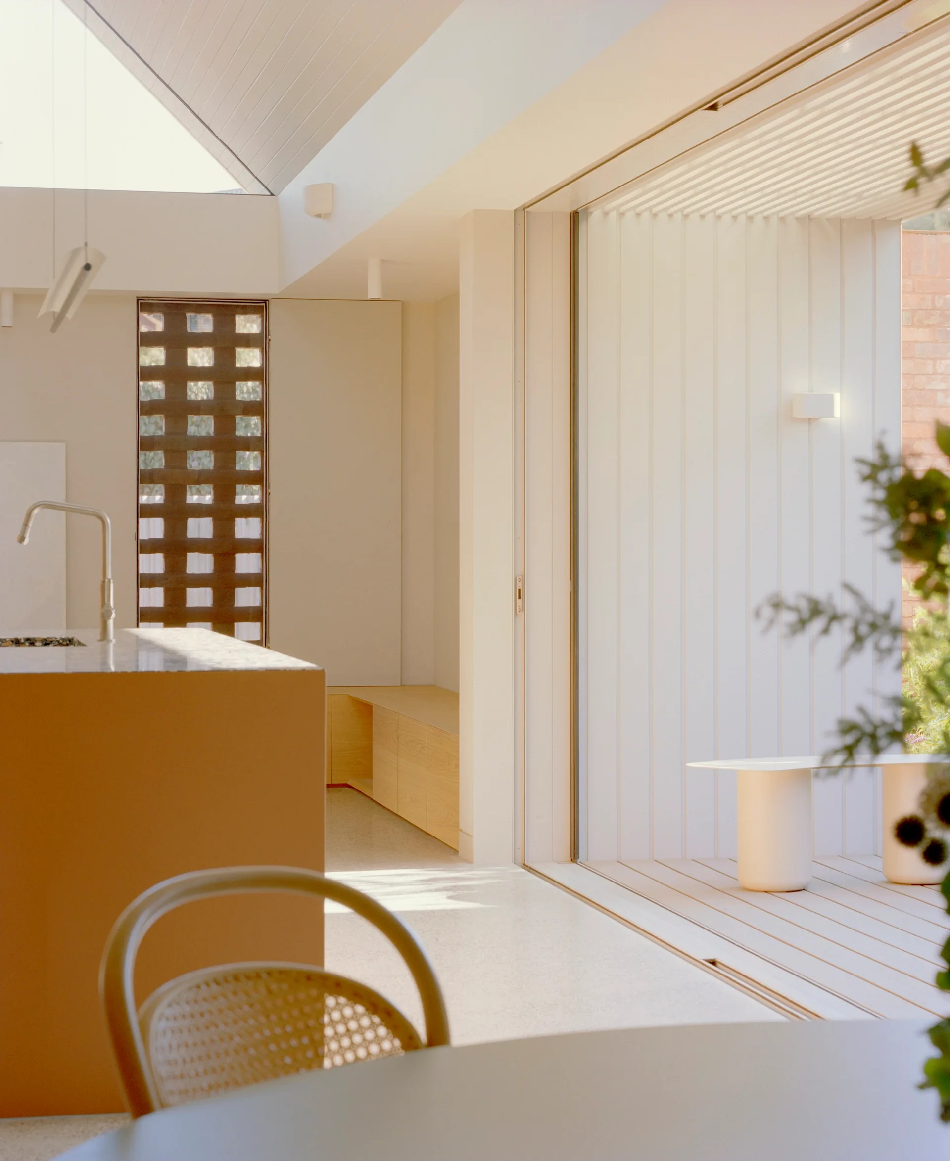 A modern minimalist kitchen features a light beige island, a low wooden storage bench, and large sliding doors that open onto a wood-planked deck.