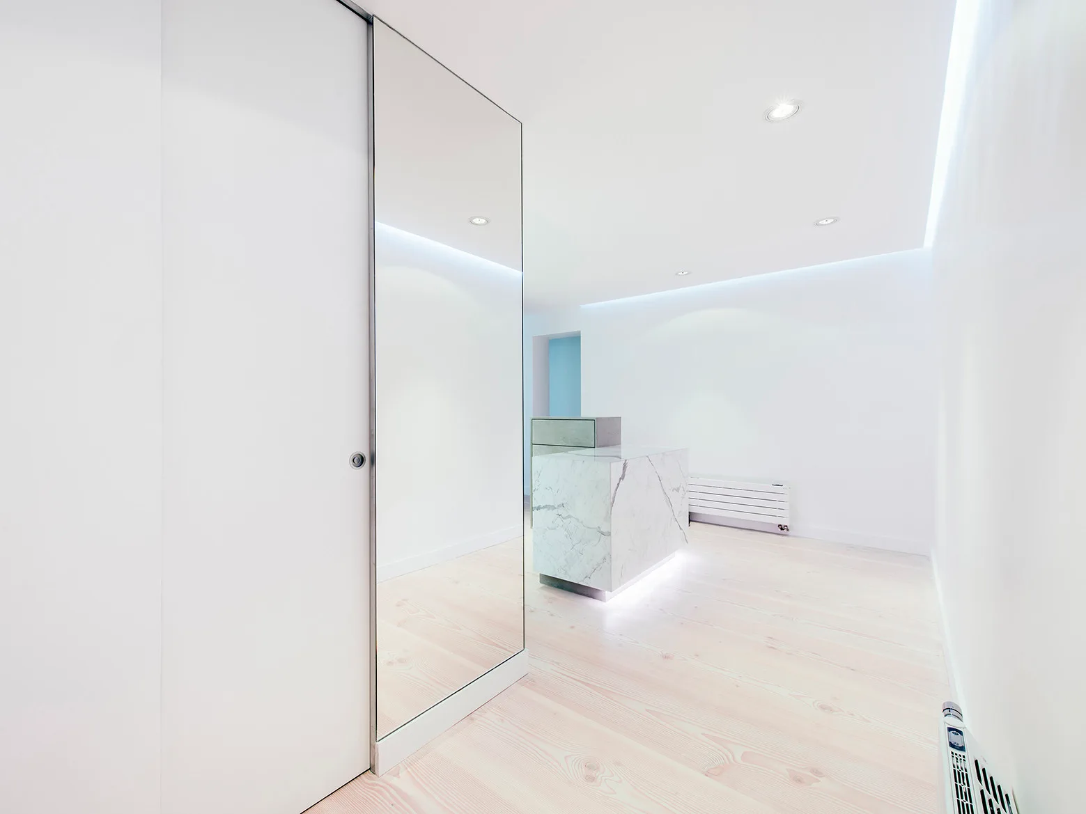A minimalist white room features a central marble island, light wood plank flooring, recessed ceiling spotlights, and a tall mirrored sliding door.