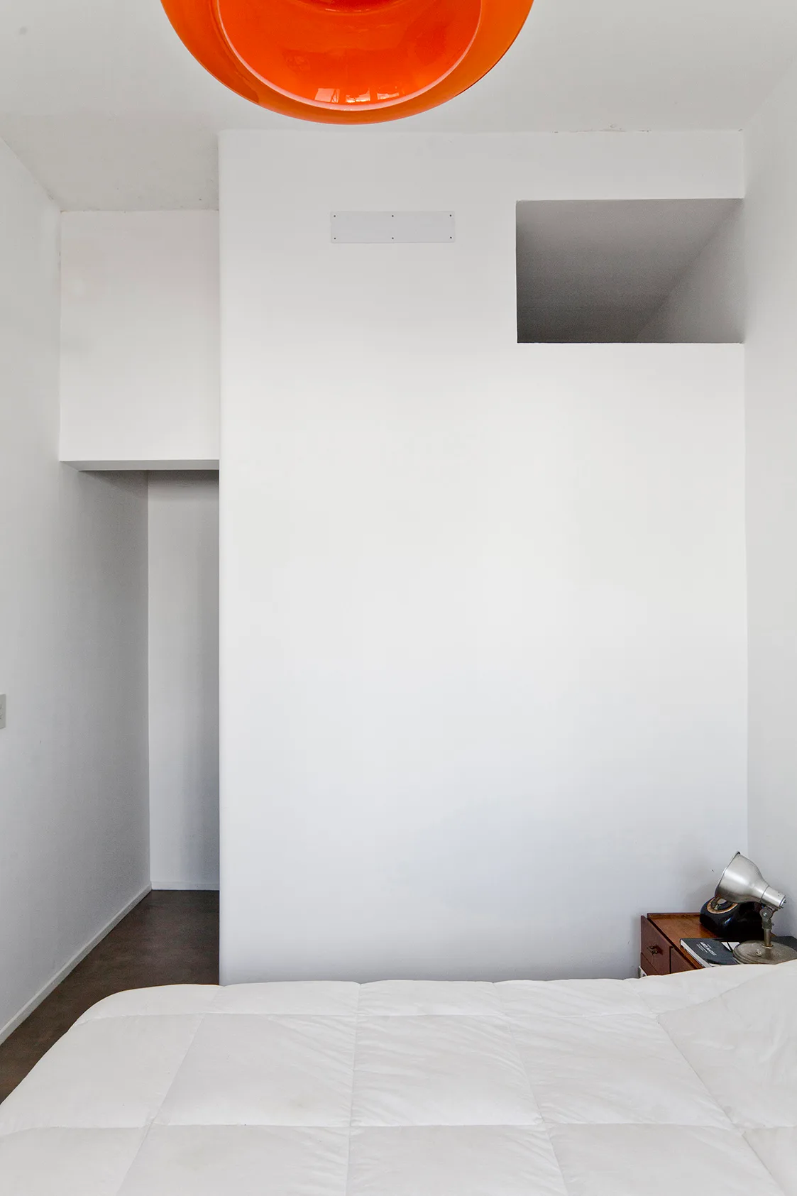 A minimalist white bedroom features architectural wall cutouts, a bed with a white duvet, a wooden nightstand, and an orange ceiling light.