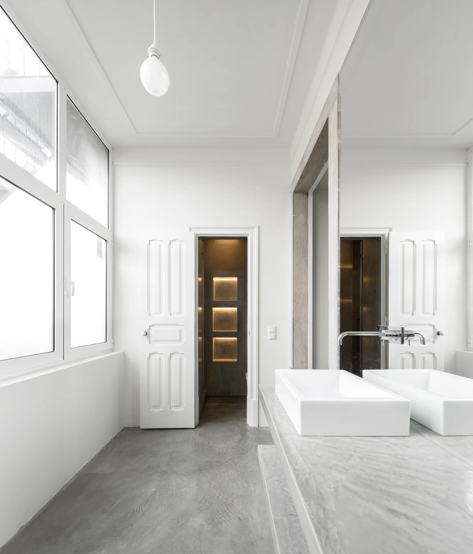 A minimalist bathroom with a concrete vanity, rectangular white vessel sink, a large mirror, and an open doorway leading to glowing wall niches.