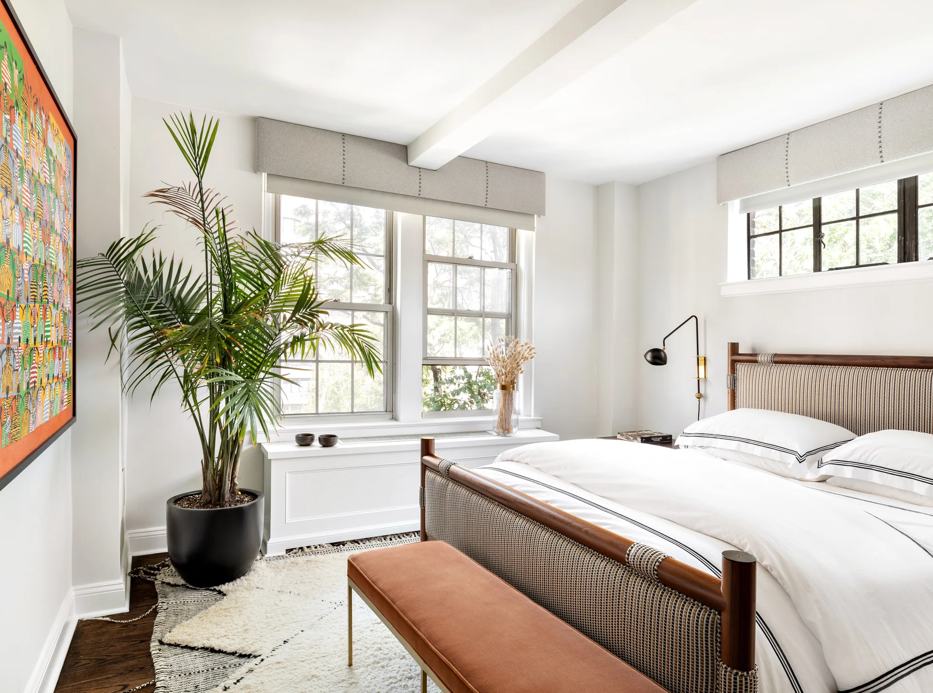 A bright white bedroom features a wood and woven bed frame, a rust-colored bench, and a large potted plant next to a window.