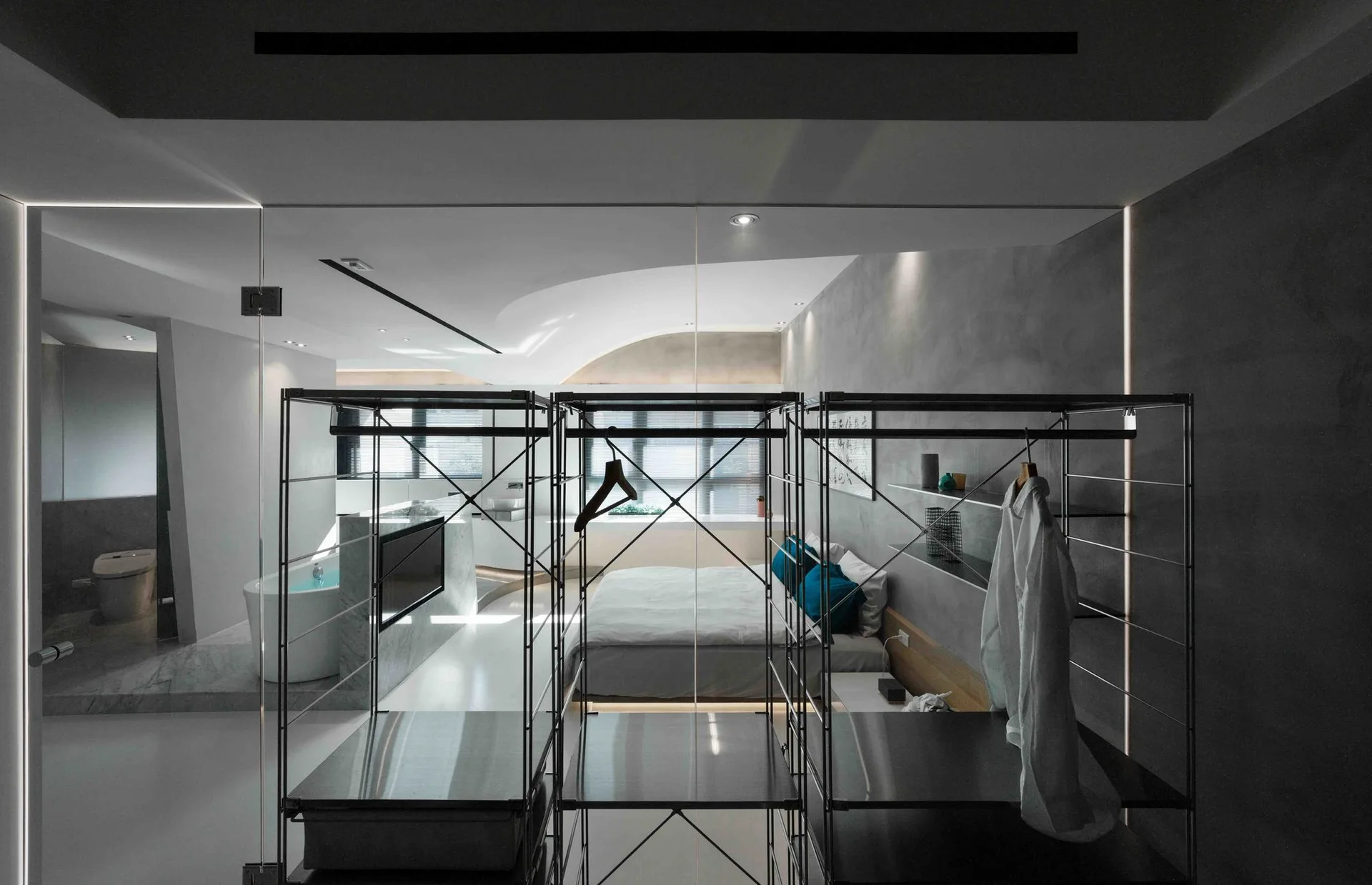 A minimalist bedroom featuring open metal closet racks behind a clear glass partition with an ensuite bathroom in the background.