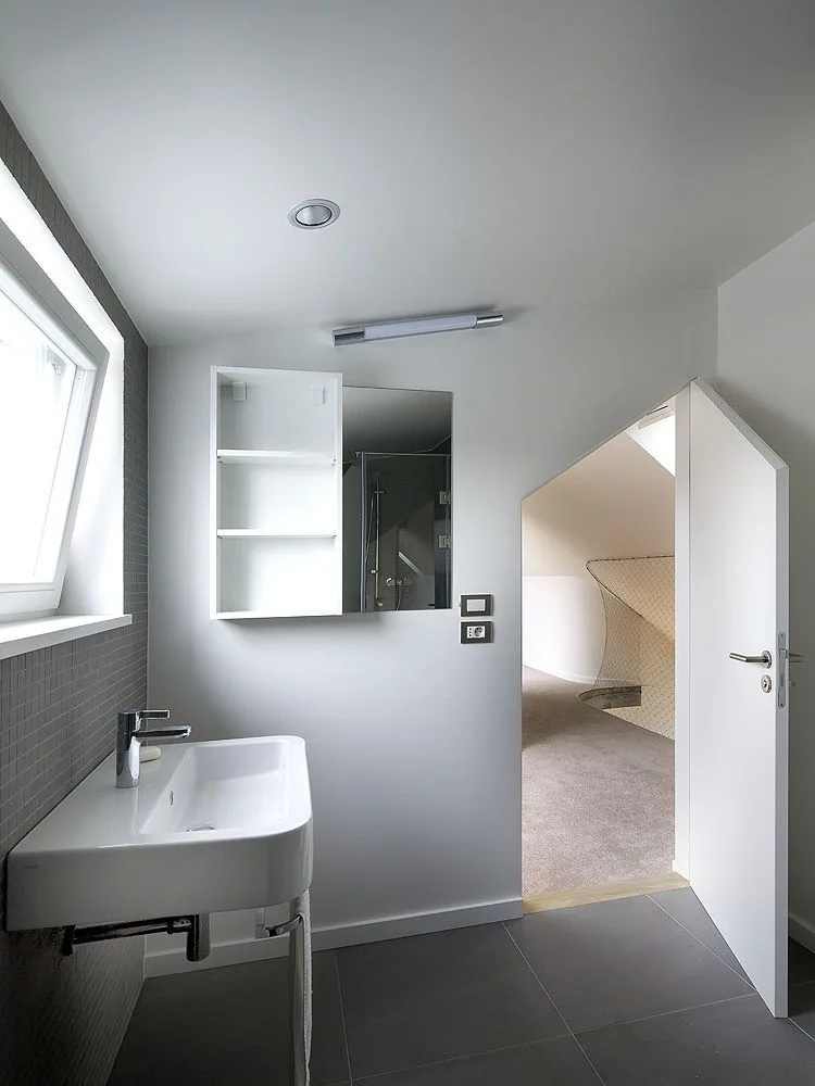 A minimalist bathroom features a white wall-mounted sink, gray mosaic tiles, and an open, angled doorway revealing a carpeted hall.
