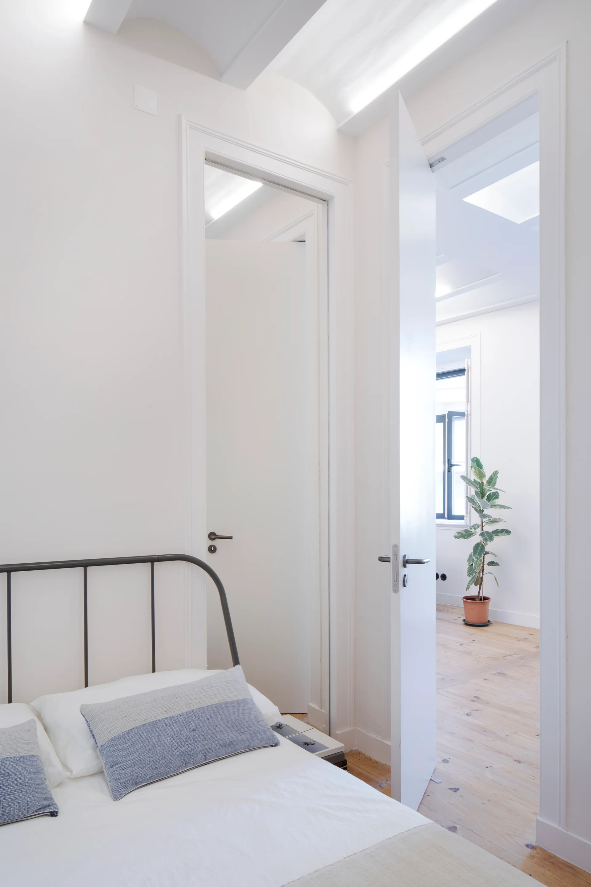 A minimalist white bedroom with a dark metal bed frame and open doors looking into an adjacent room with light wood floors and a potted plant.