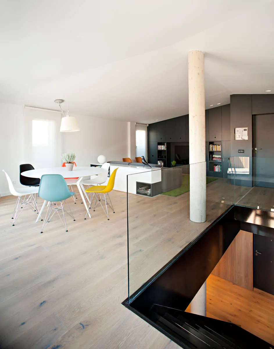 An open-plan dining room with a white circular table, multicolored chairs, a central concrete column, and a staircase with a glass railing.