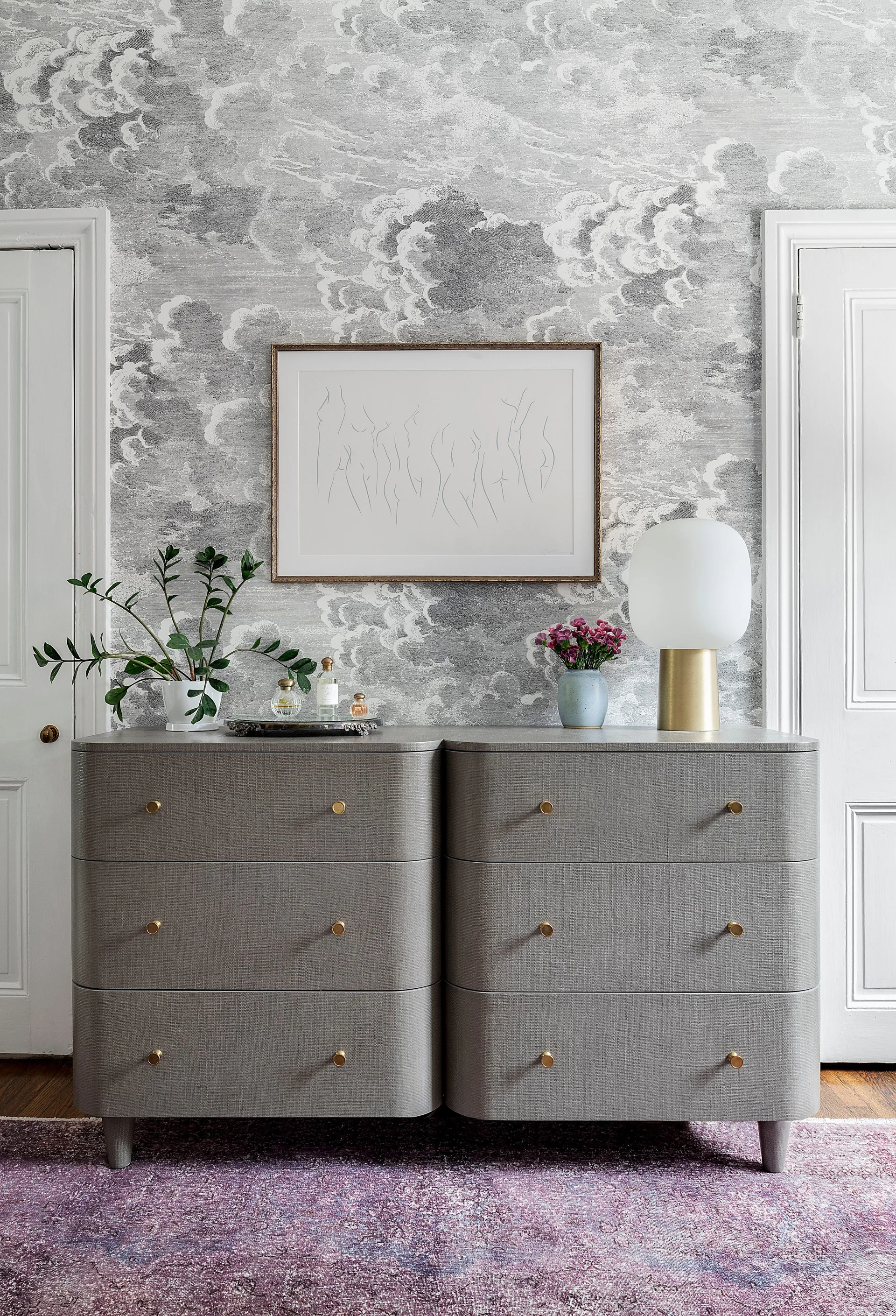 A textured grey dresser with rounded corners and gold knobs sits against grey cloud-patterned wallpaper between two white doors.