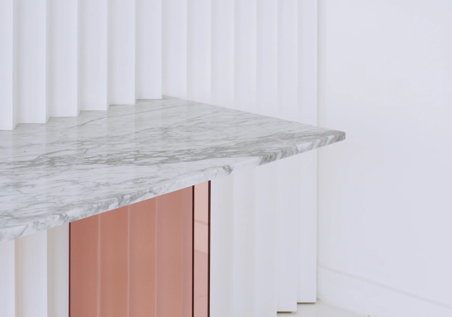 A white veined marble tabletop rests against a fluted white wall with a reflective rose gold metallic base beneath it.