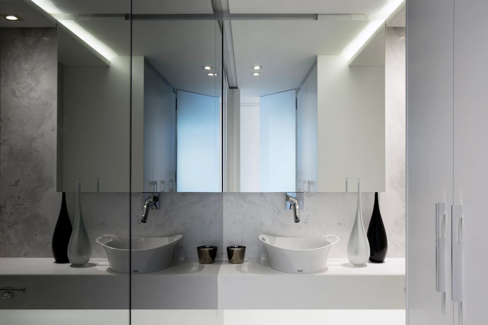 A minimalist white bathroom vanity features two ceramic vessel sinks, wall-mounted chrome faucets, a veined marble backsplash, and a large backlit mirror.