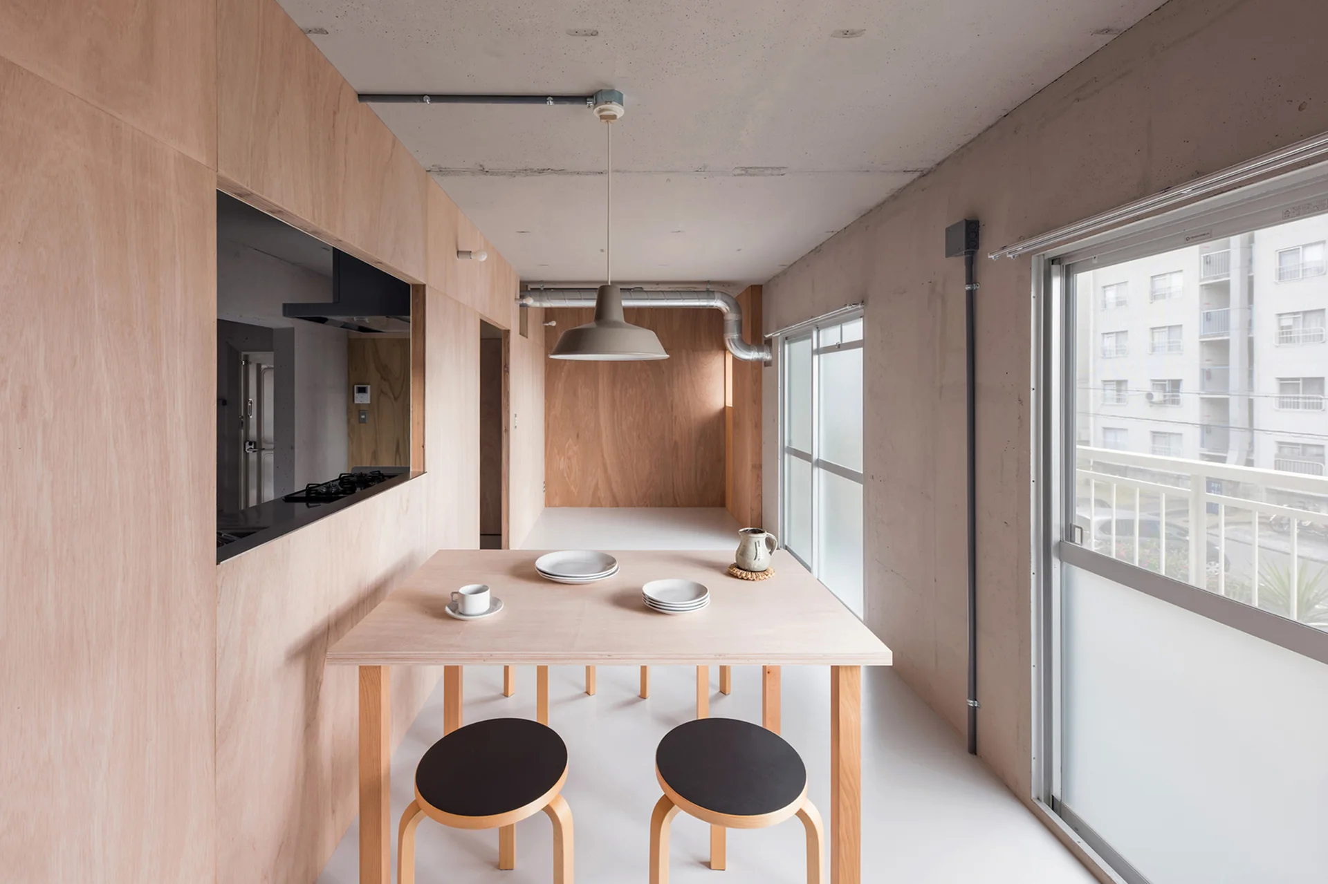 A minimalist dining area features a light wood table with two black-topped stools set against plywood-clad walls and a raw concrete ceiling.
