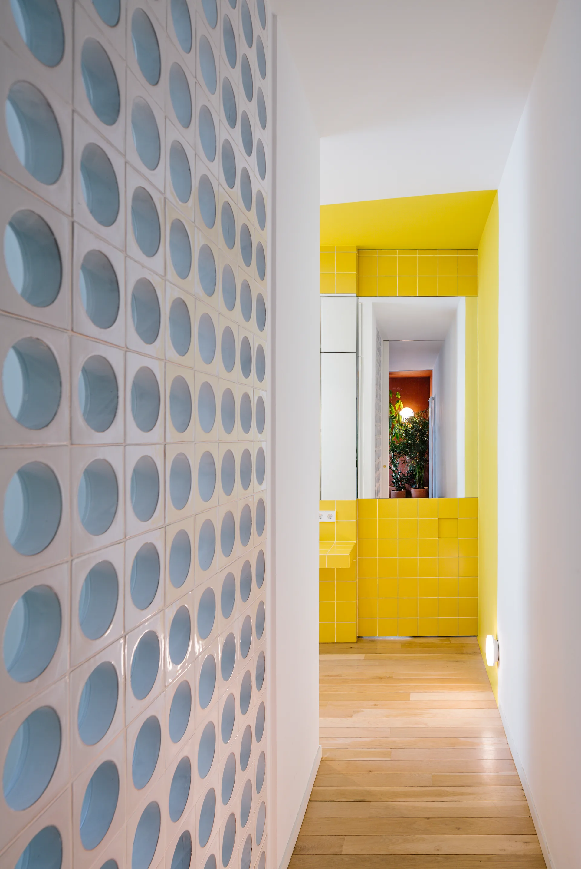 A modern hallway features a white wall with repeating circular cutouts, hardwood flooring, and a bright yellow tiled bathroom visible at the end.
