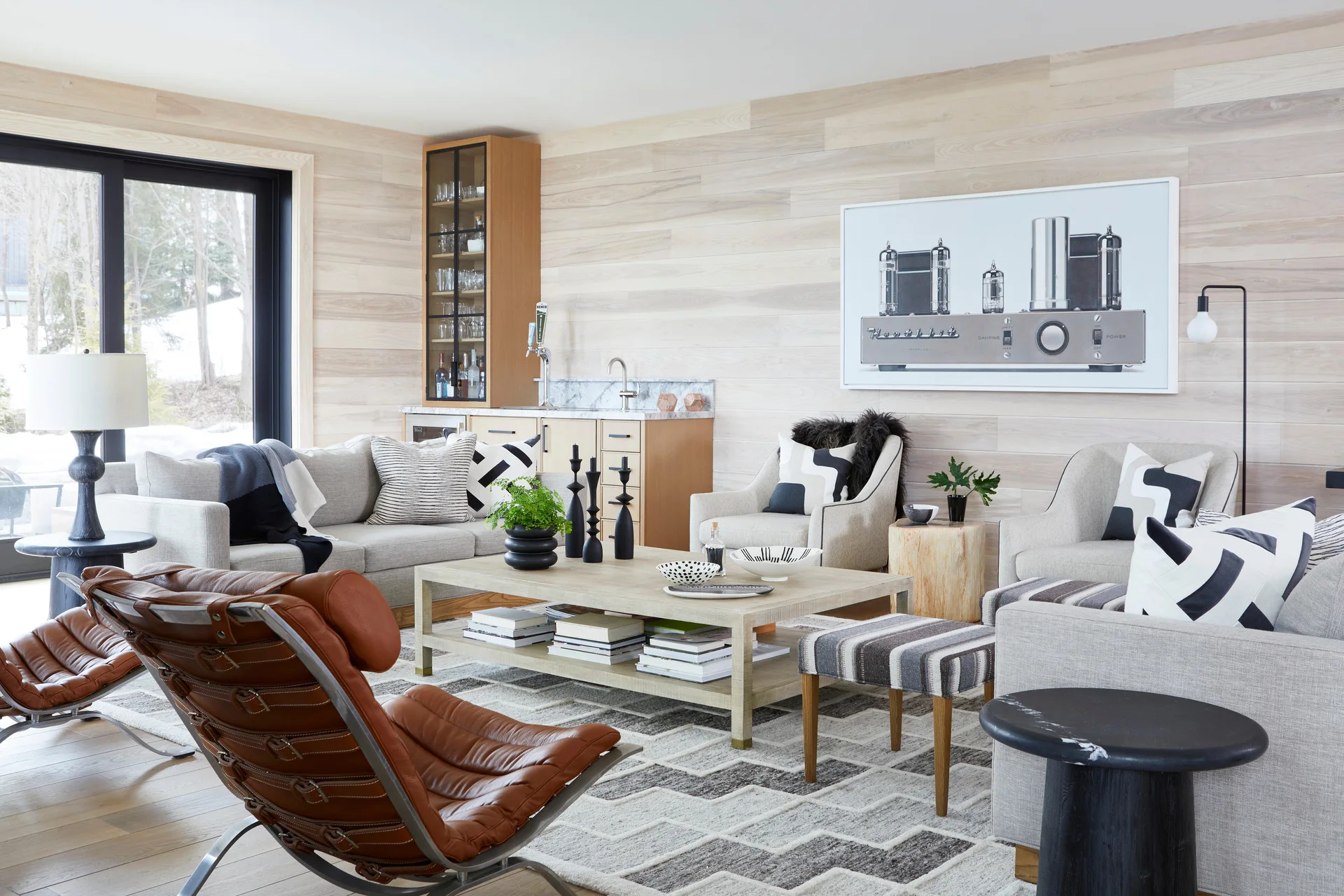 A bright living room features pale horizontal wood wall paneling, light grey sofas with graphic pillows, a leather lounge chair, and a wet bar.