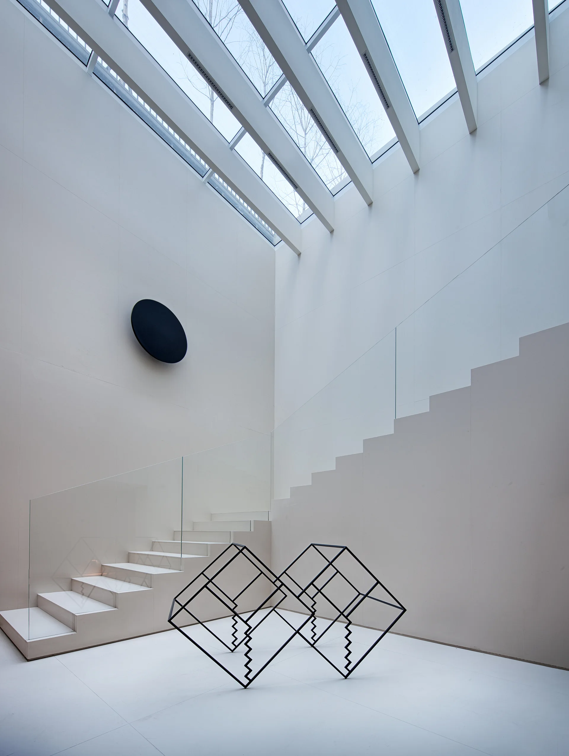 A minimalist white atrium featuring a floating staircase with glass railings, a black geometric floor sculpture, and a large grid-pattern skylight ceiling.