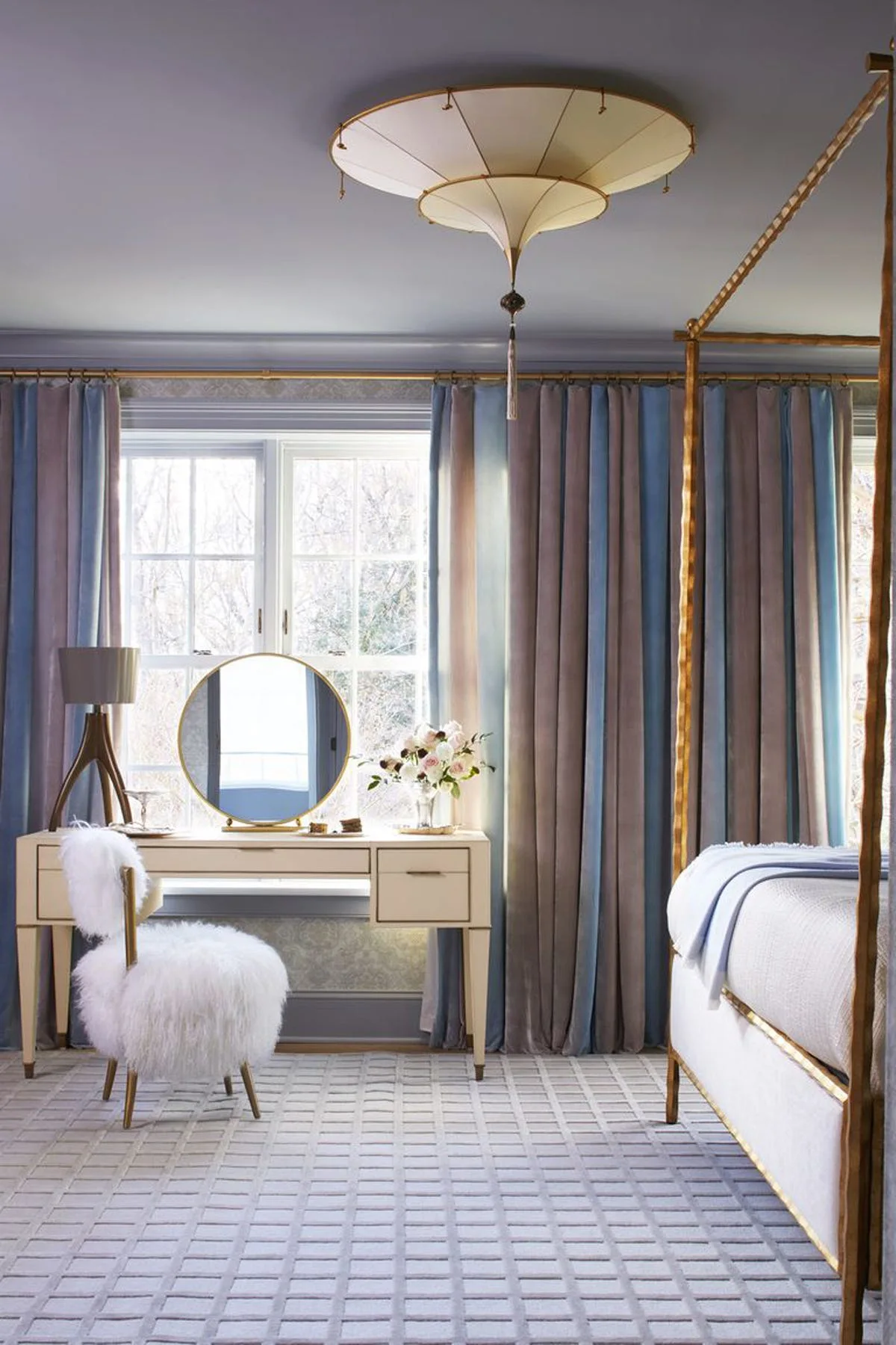 A bedroom dressing area features a cream vanity desk with a round mirror, a white furry chair, and long blue drapes beside a gilded bed frame.