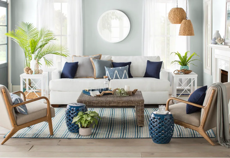 A coastal living room features a white sofa, a woven wicker coffee table, blue ceramic garden stools, and a striped blue and white rug.