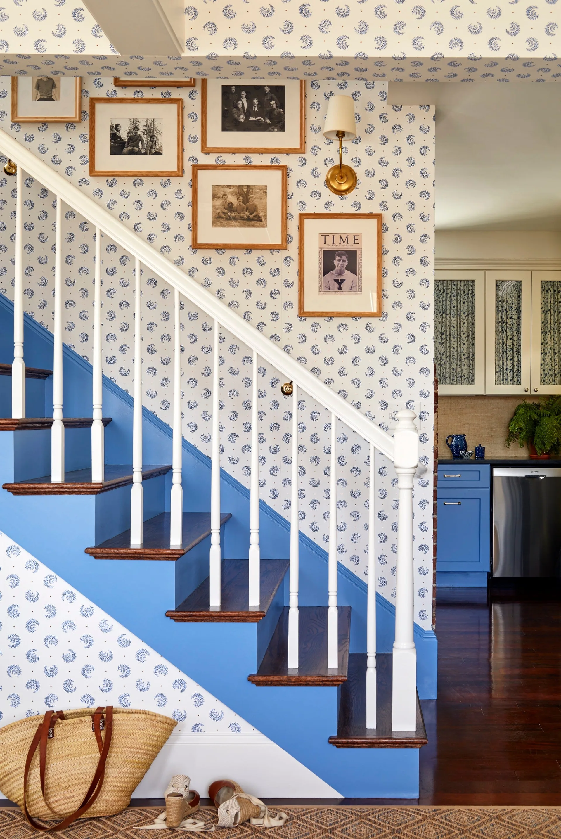 A staircase with blue painted risers and white spindles features blue and white patterned wallpaper, dark wood treads, and a gallery wall.